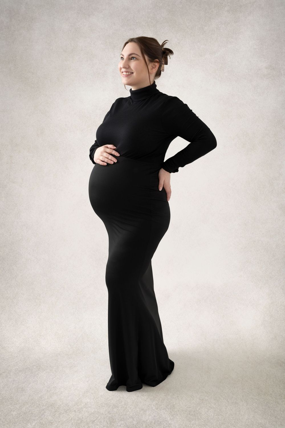 Smiling pregnant woman in elegant black turtleneck maxi dress, cradling baby bump, studio portrait.