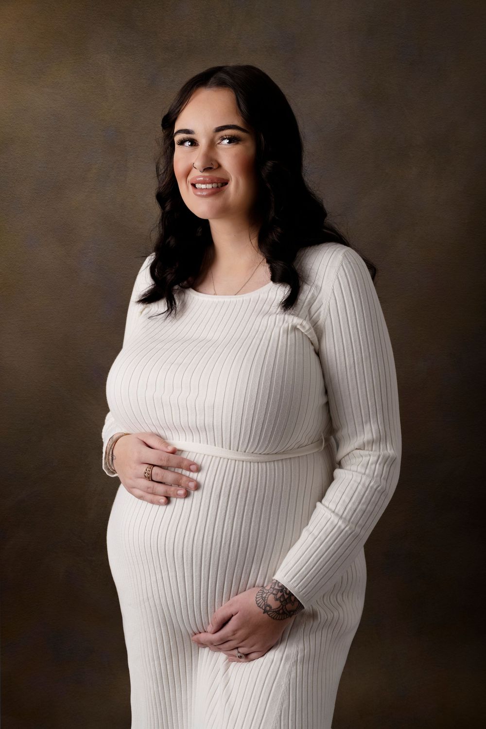 Expectant mother in white ribbed dress posing against dark backdrop.