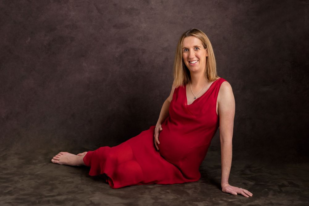 A pregnant mother in a sleeveless red dress poses gracefully on the floor on a dark studio backdrop.
