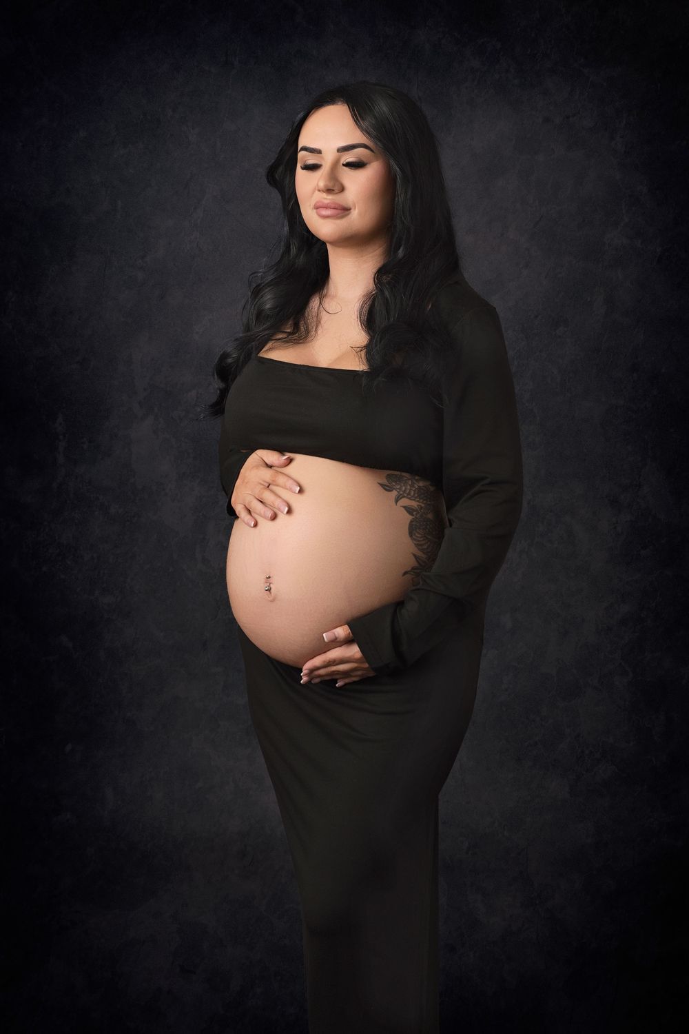 Dramatic maternity portrait in black dress against dark studio backdrop.