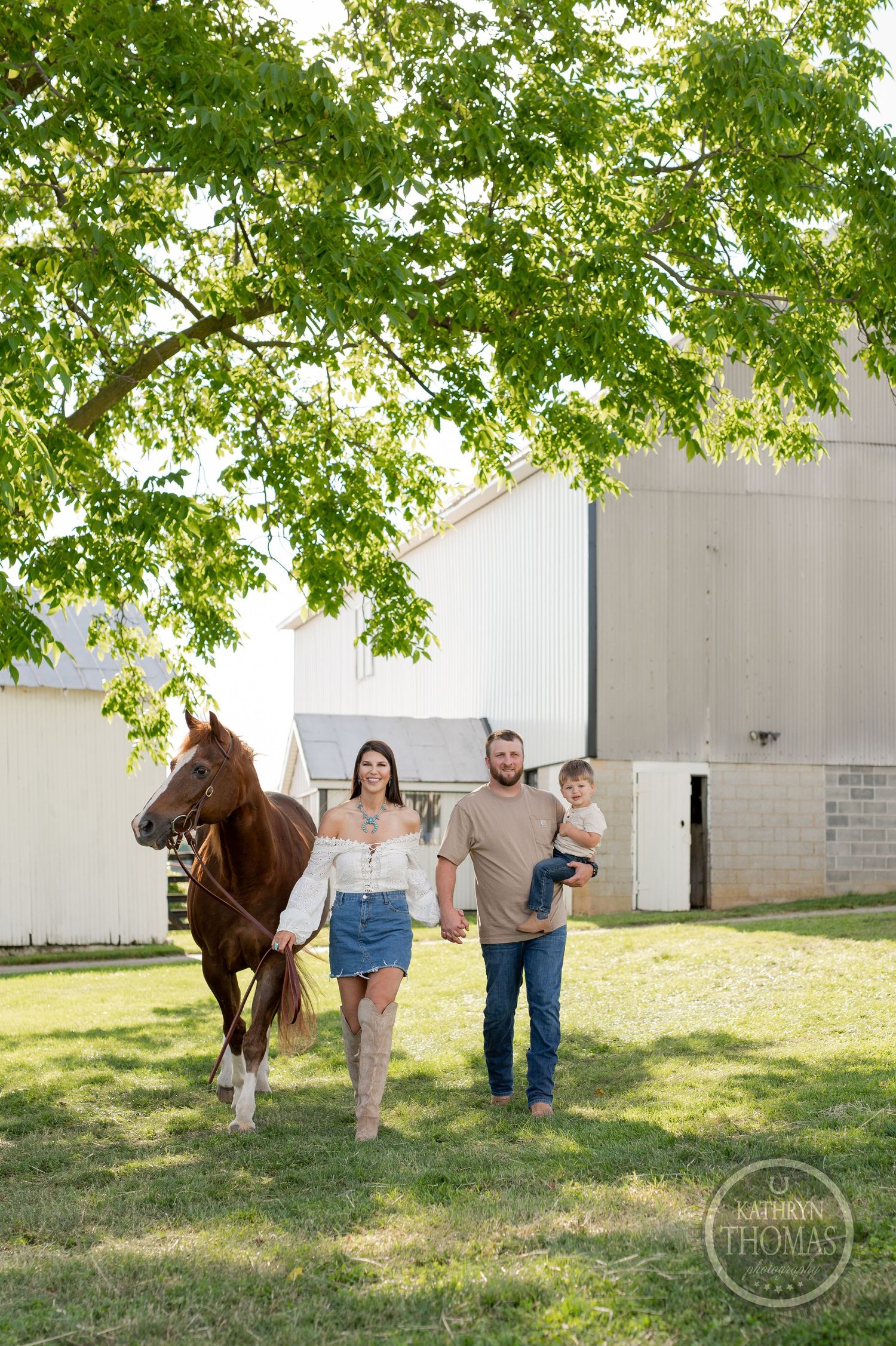 Family and horse walking together near a white barn under green summer trees.