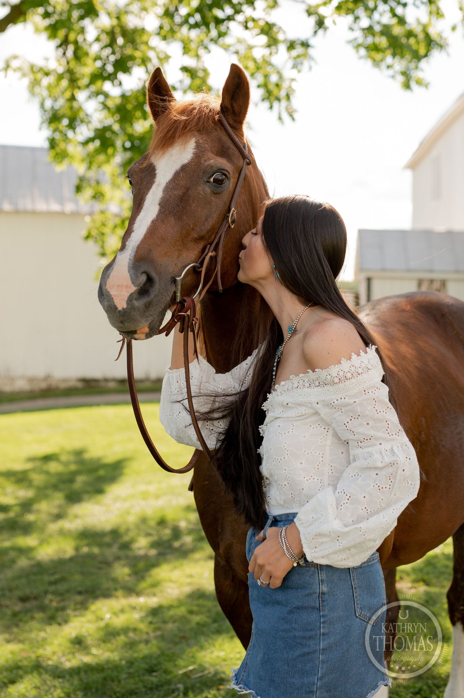 A sweet moment between a chestnut horse and someone in a white peasant top near farm buildings.