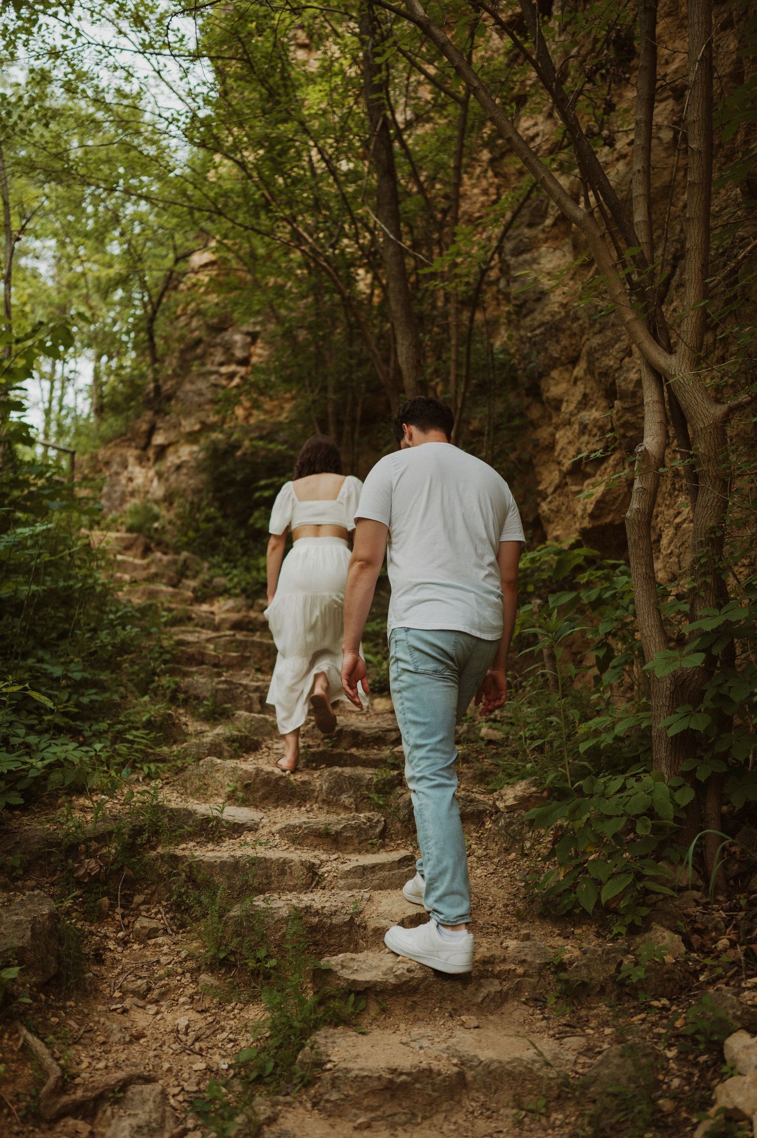 A couple climb stone steps together on a wooded hiking trail surrounded by lush greenery.