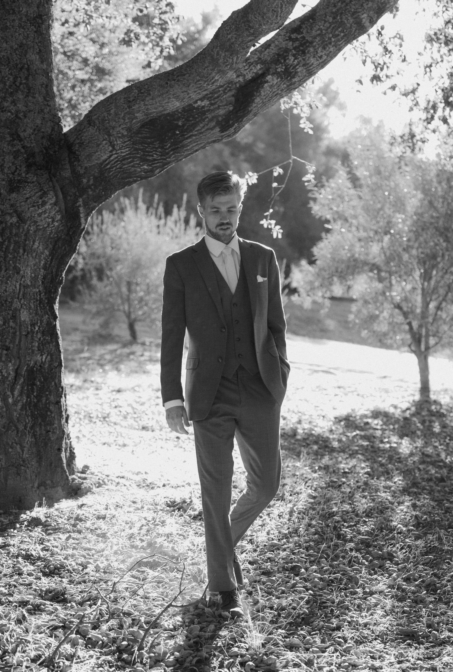 Black and white portrait of someone in a dark suit leaning against a tree trunk in a sunlit outdoor setting.