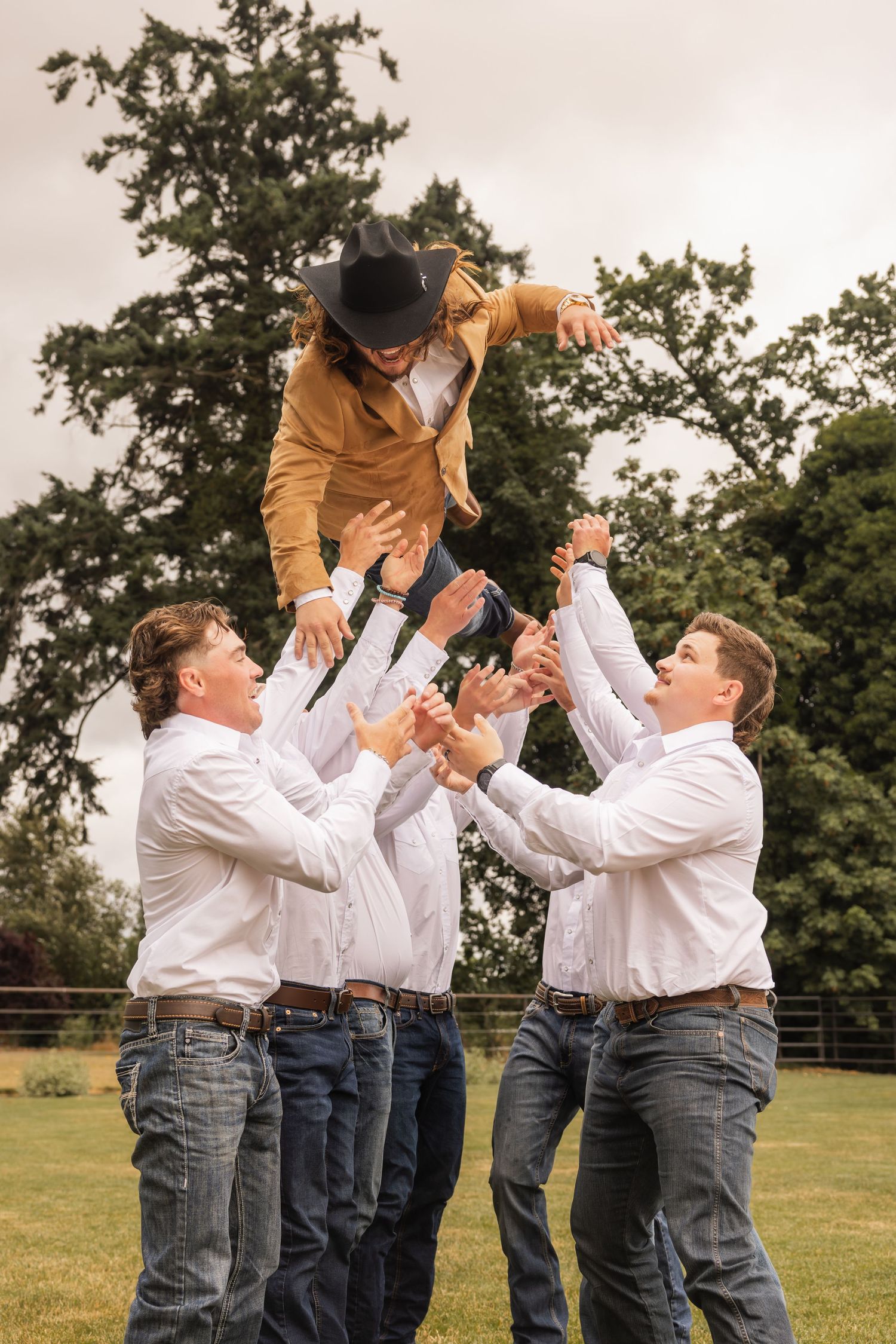 Groomsmen in white shirts and jeans cheerfully toss someone wearing a mustard jacket and black hat in outdoor celebration.