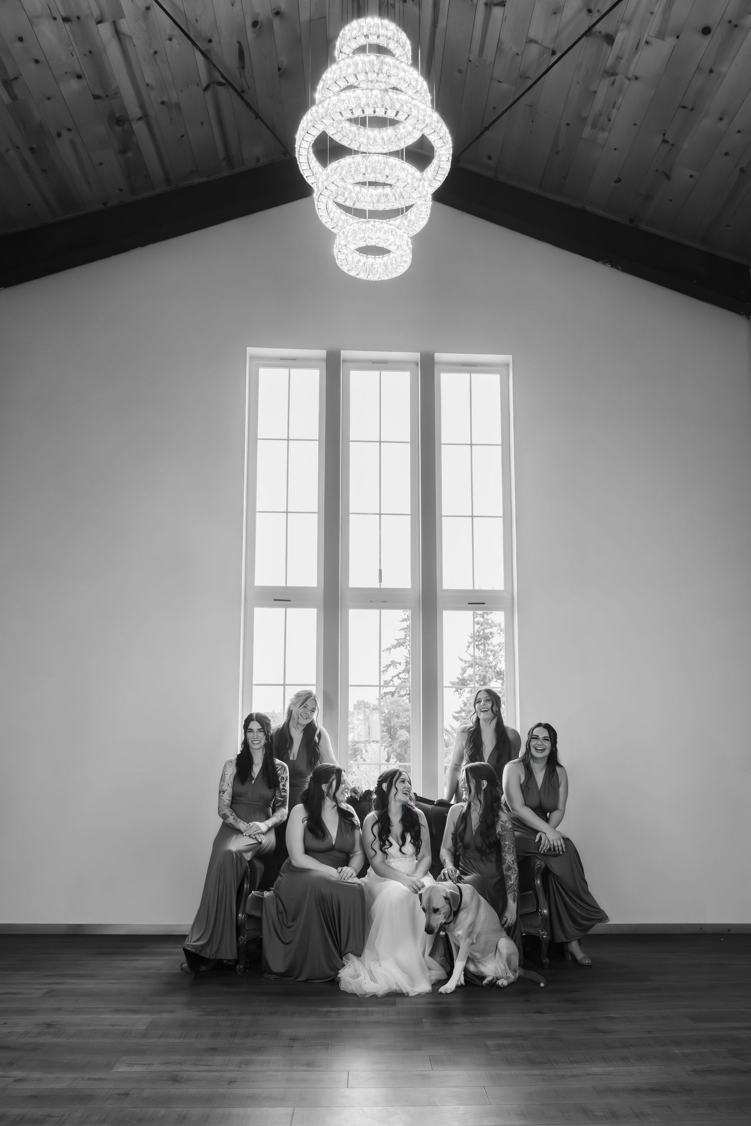 Black and white photo of women in formal dresses gathered under a crystal chandelier in an elegant room with tall windows.