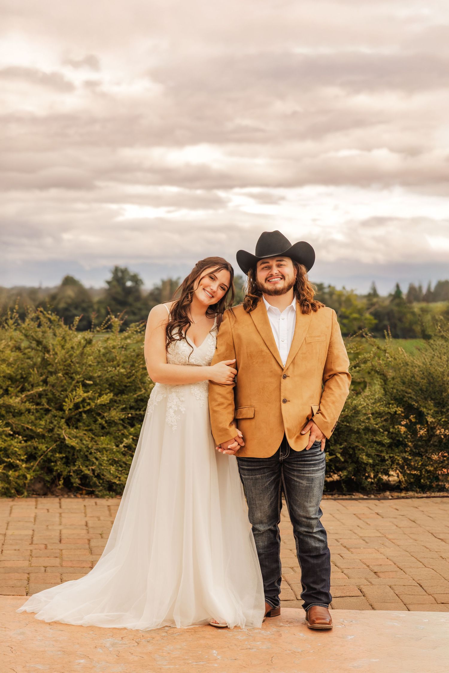 A couple poses outdoors for their wedding photo with rustic country style on a cloudy day in front of green foliage.