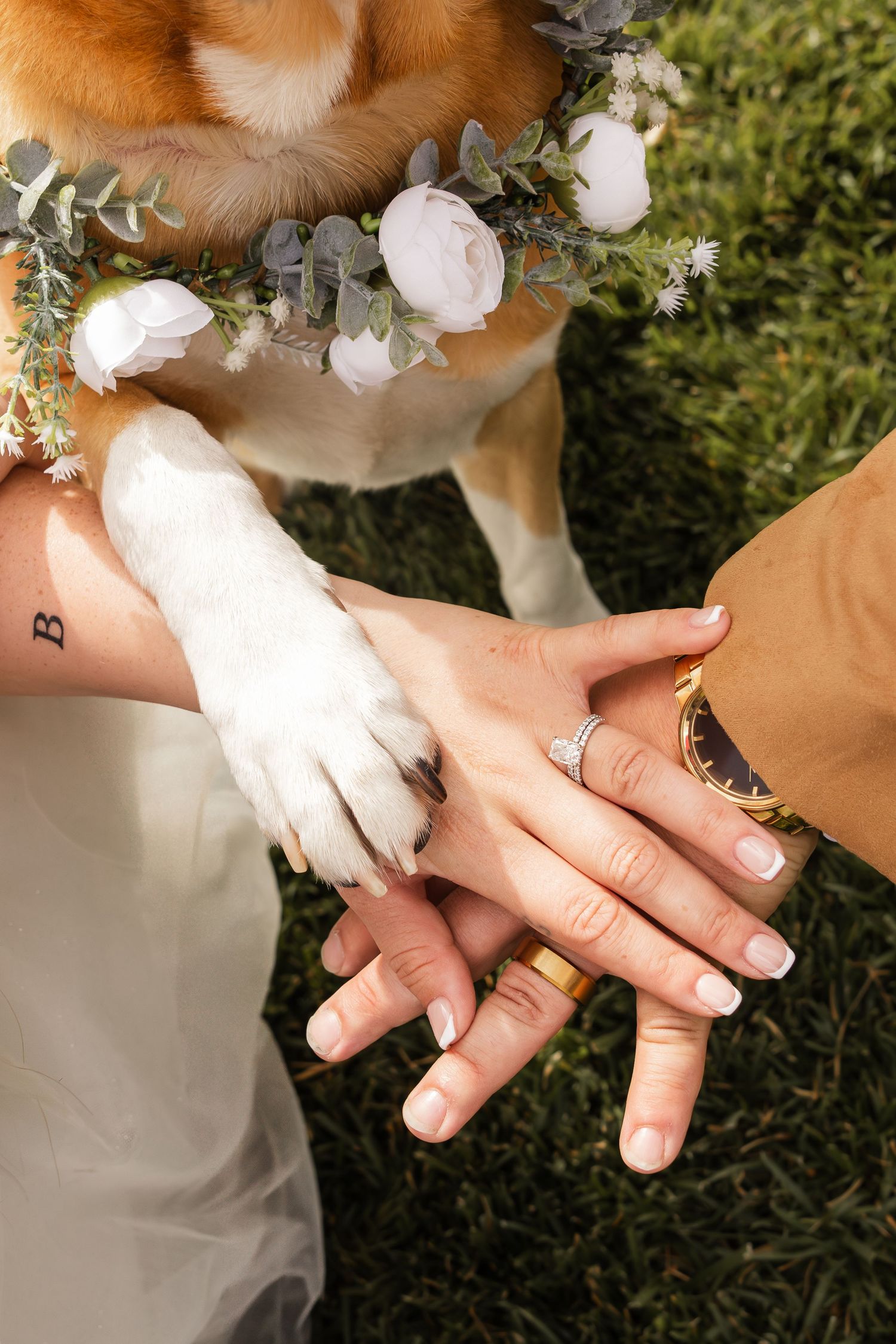 Wedding ring detail shot shows hands exchanging bands with delicate white flowers and greenery in the background.