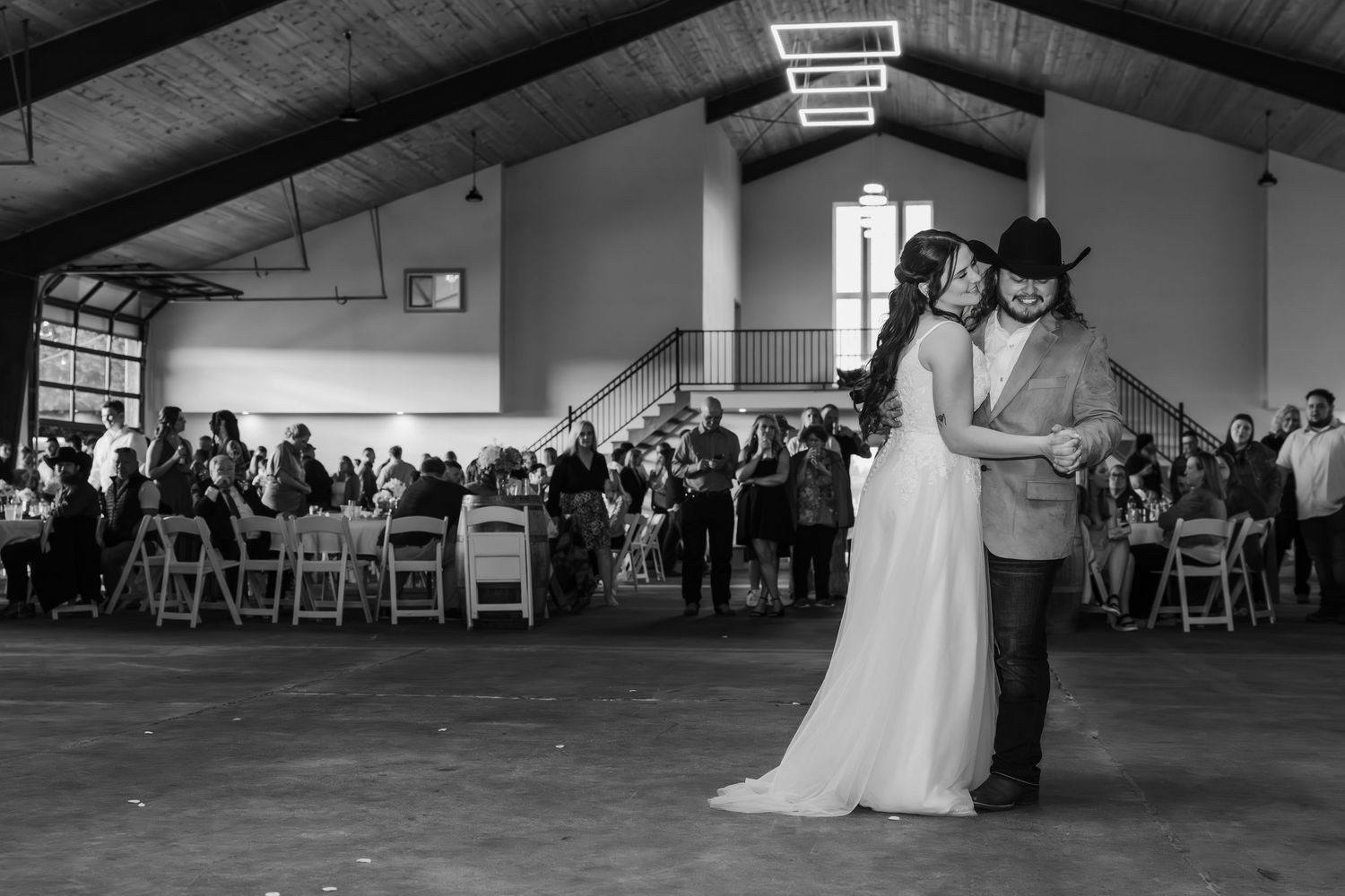 A couple shares their first dance at a wedding reception in a large venue with guests seated at tables in black and white.
