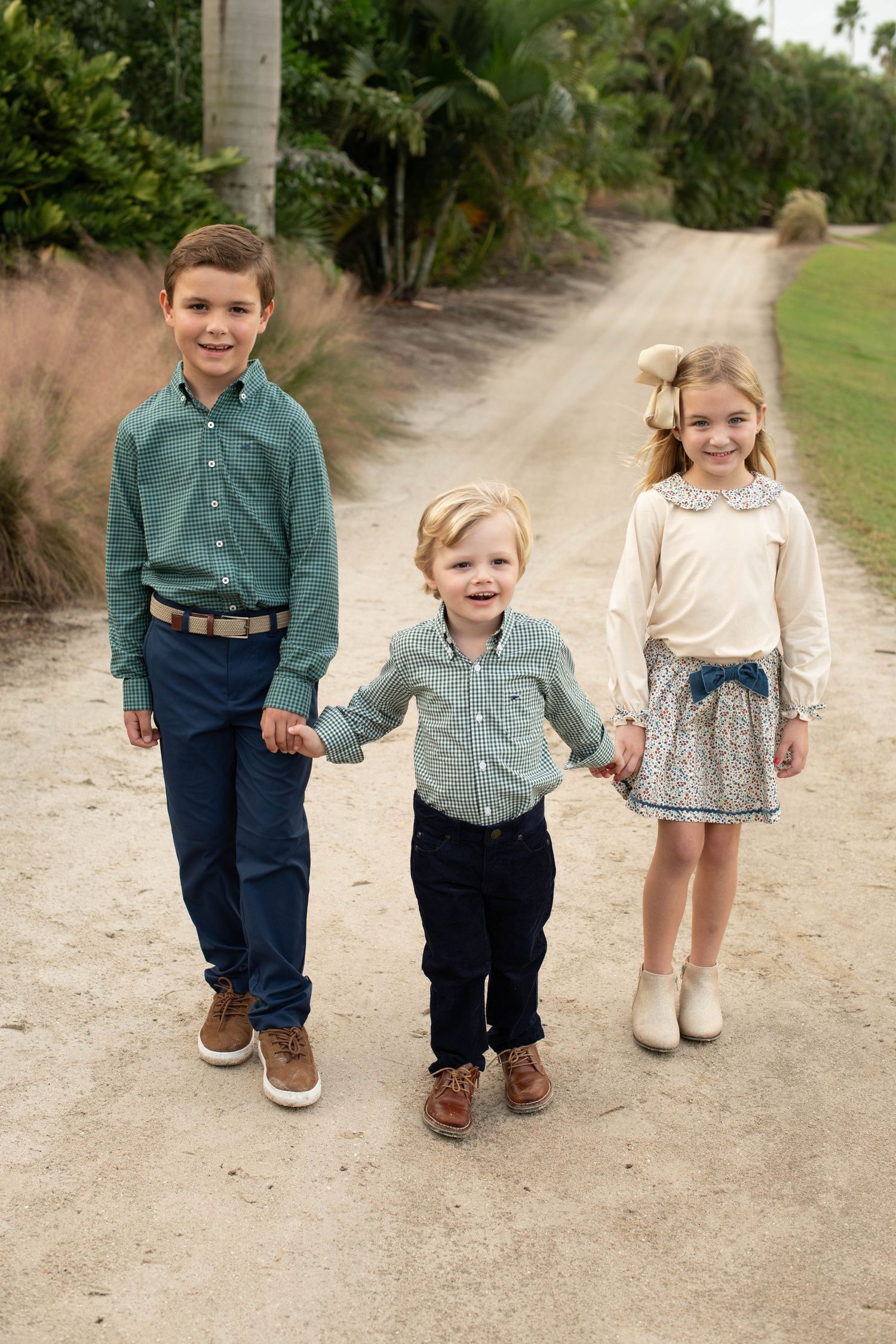 Three young children hold hands while walking down a sandy path bordered by palm trees.