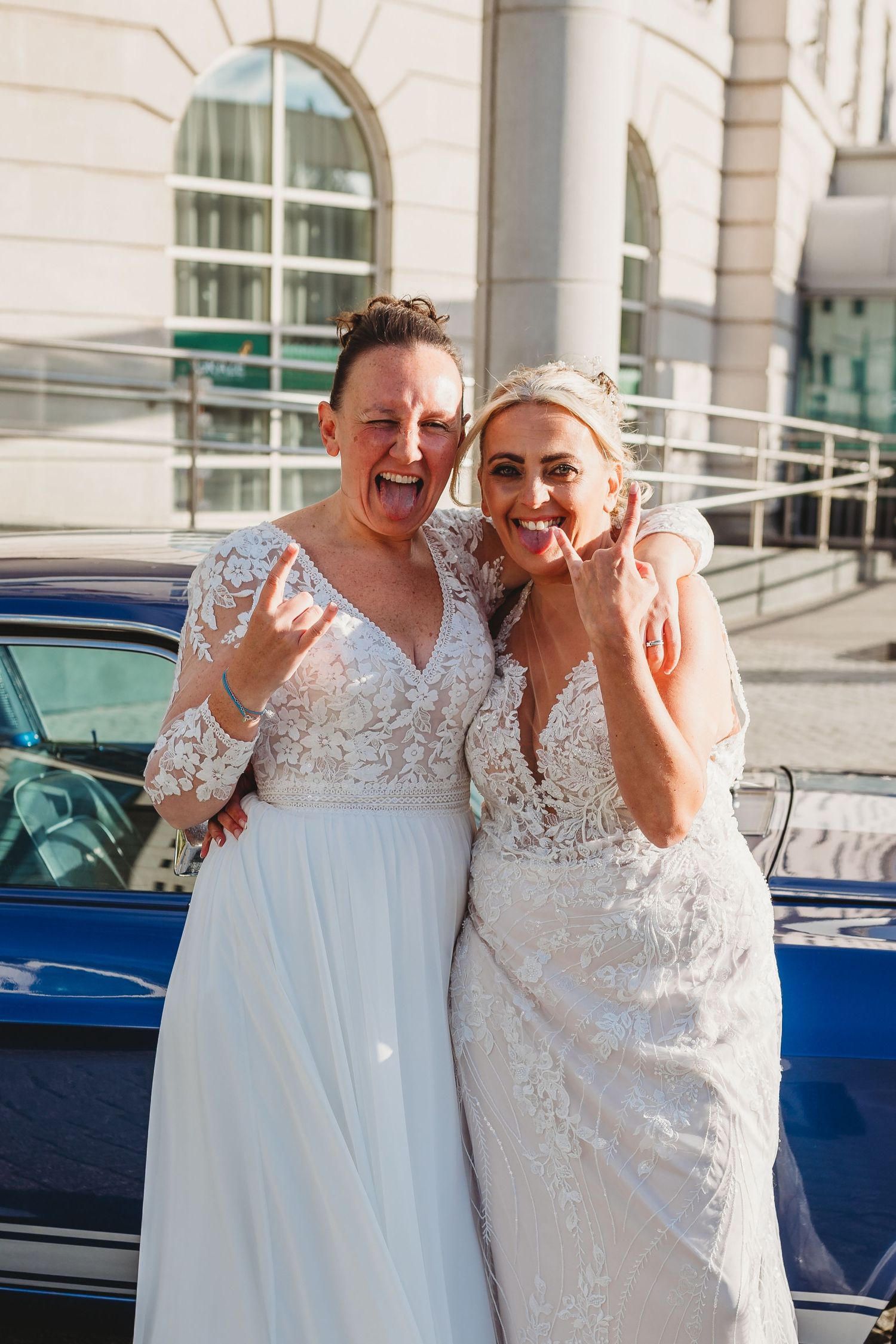 Two people in white lace wedding dresses share a joyful moment outdoors.