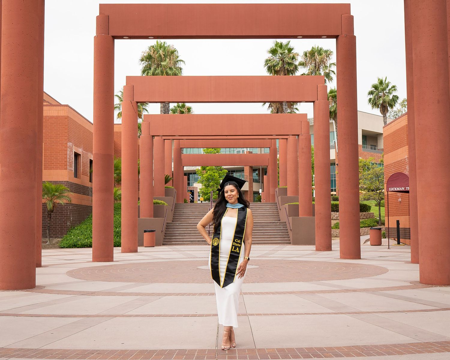 Graduate in white dress and Cal State LA stole poses under large red campus arches.