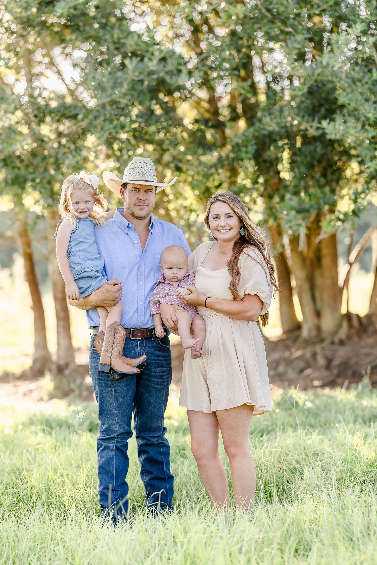 A family stands together in a sunlit grassy field with trees providing a natural backdrop in soft golden light.