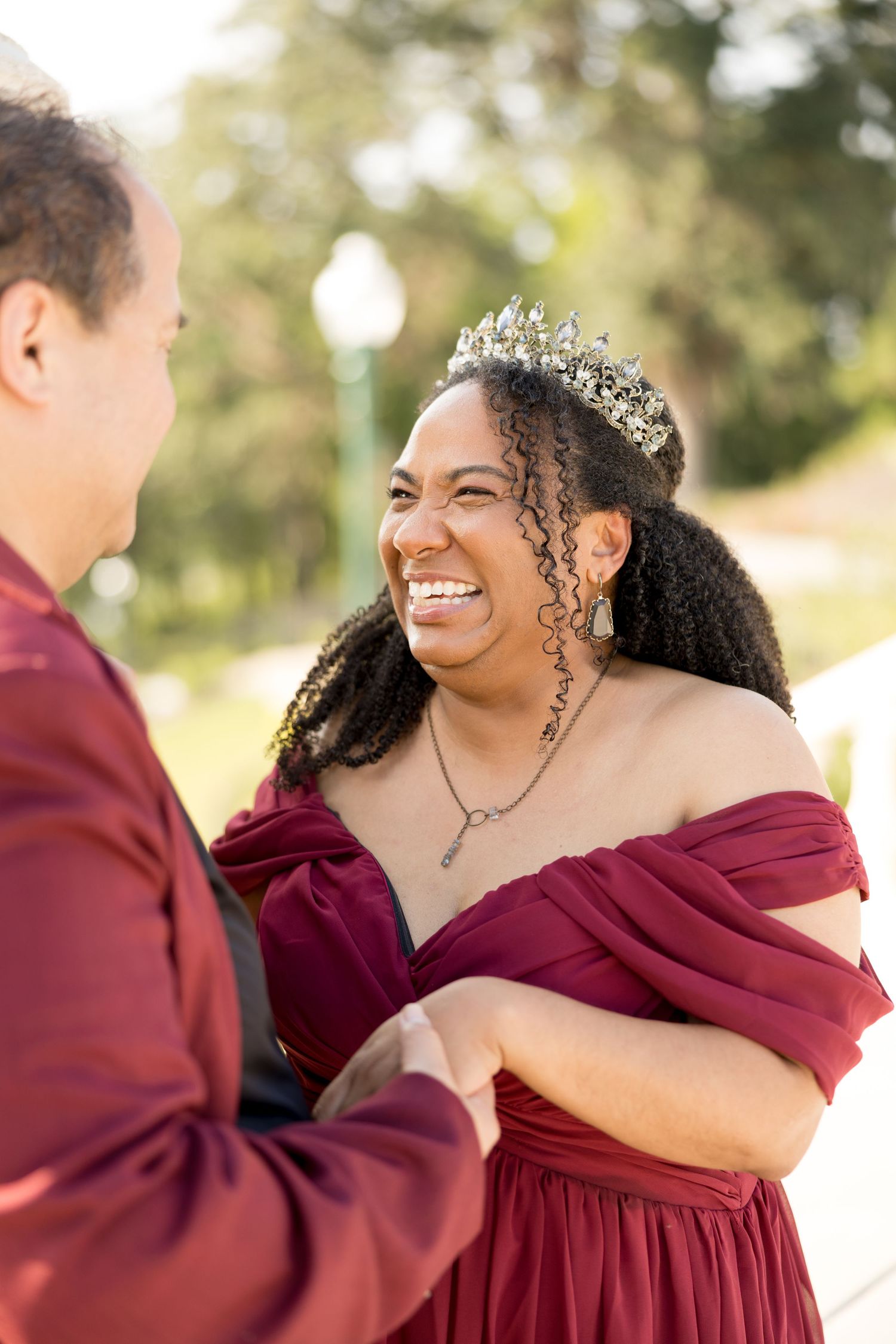 A couple in burgundy wedding attire sharing joyful laughter during an outdoor photo session.