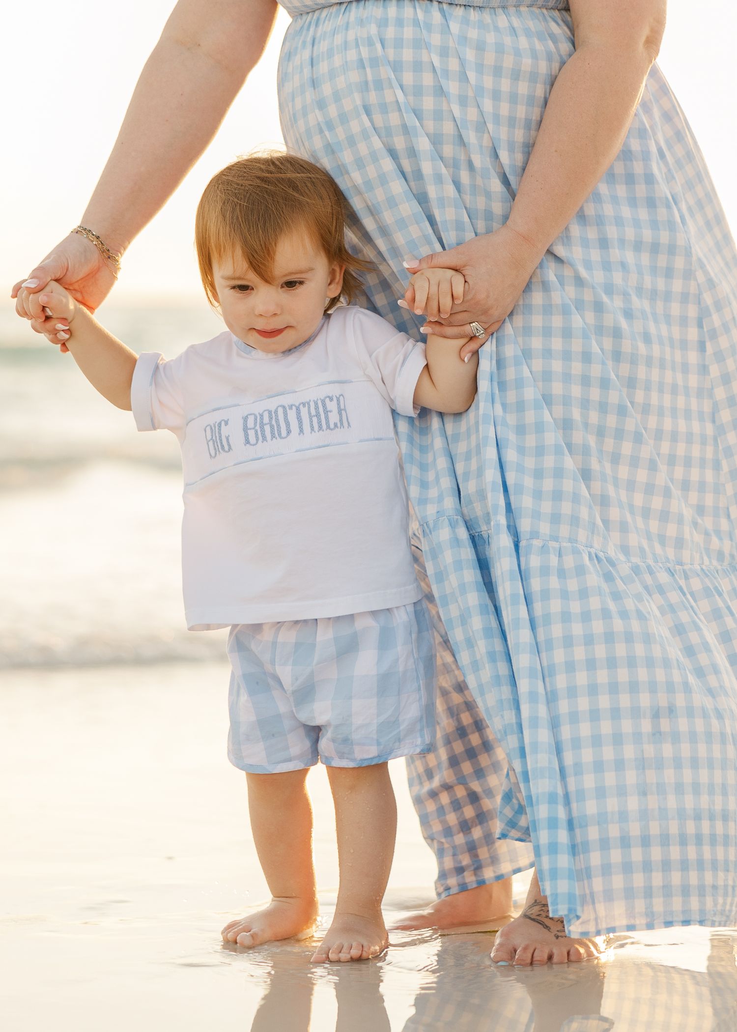 Parent and small child in matching blue gingham outfits hold hands while standing barefoot on a sandy beach.