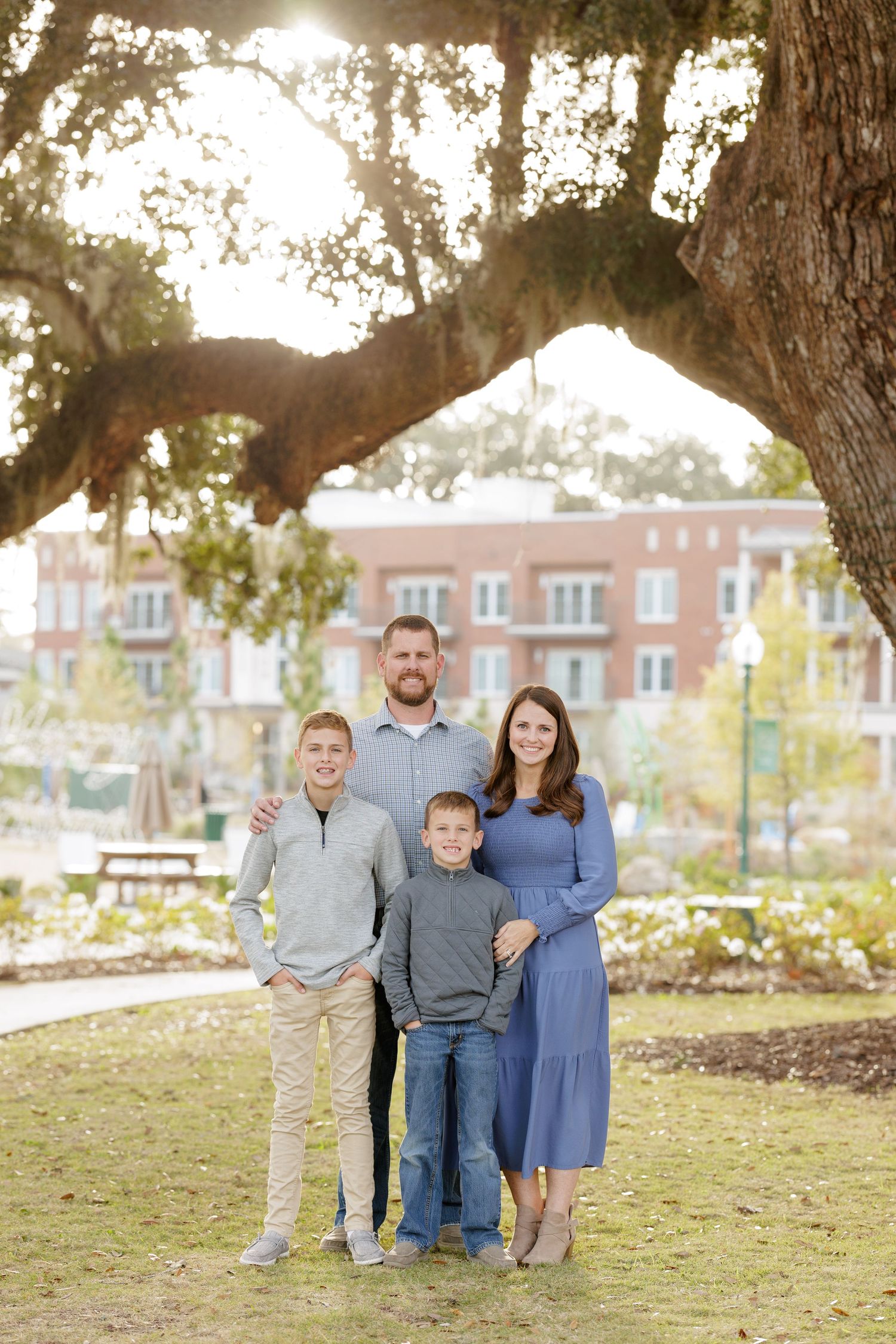 A family of four poses outdoors under large oak trees during a golden sunset photo session.