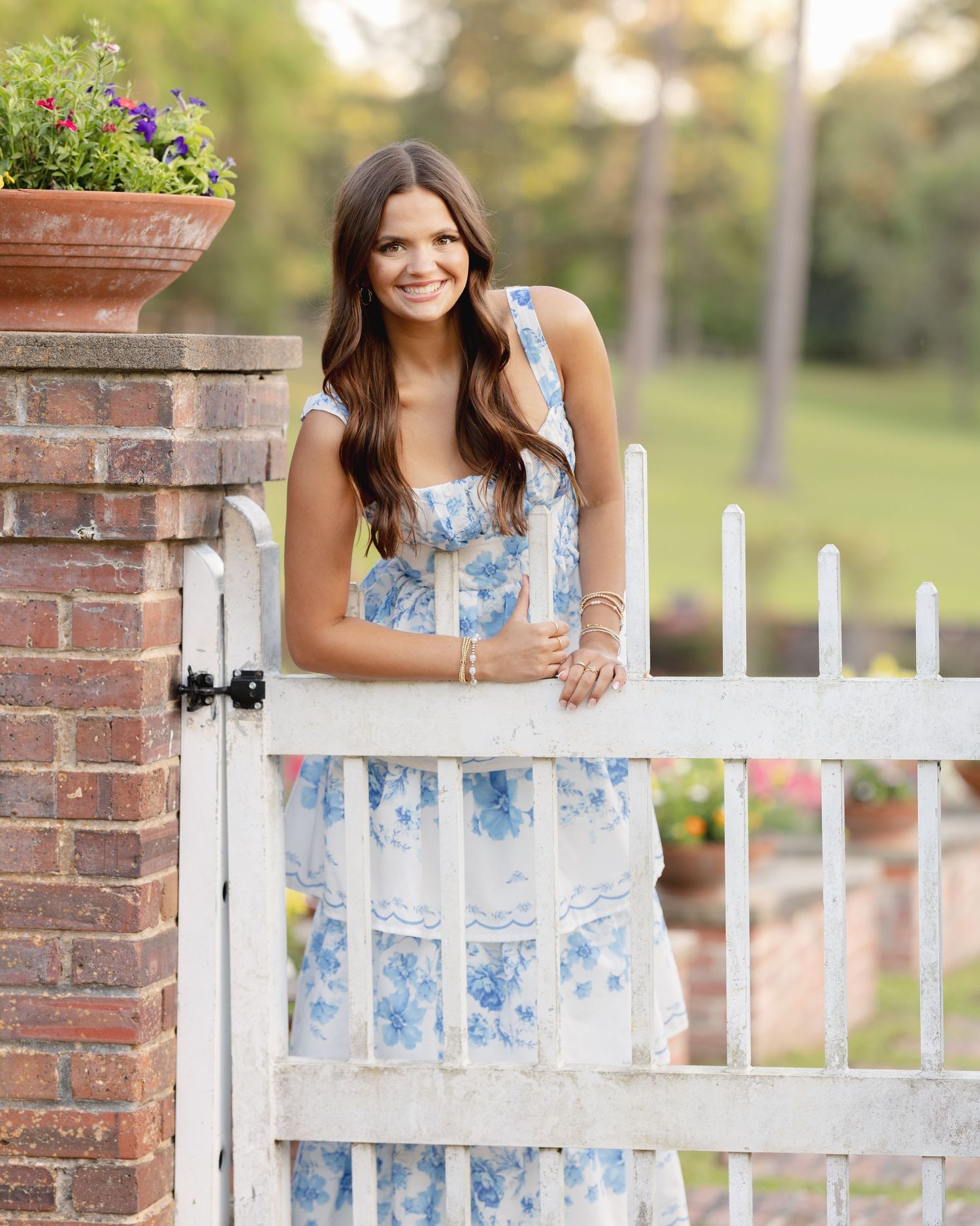 Young woman in a blue floral dress poses by a white garden gate and brick pillar with potted flowers.