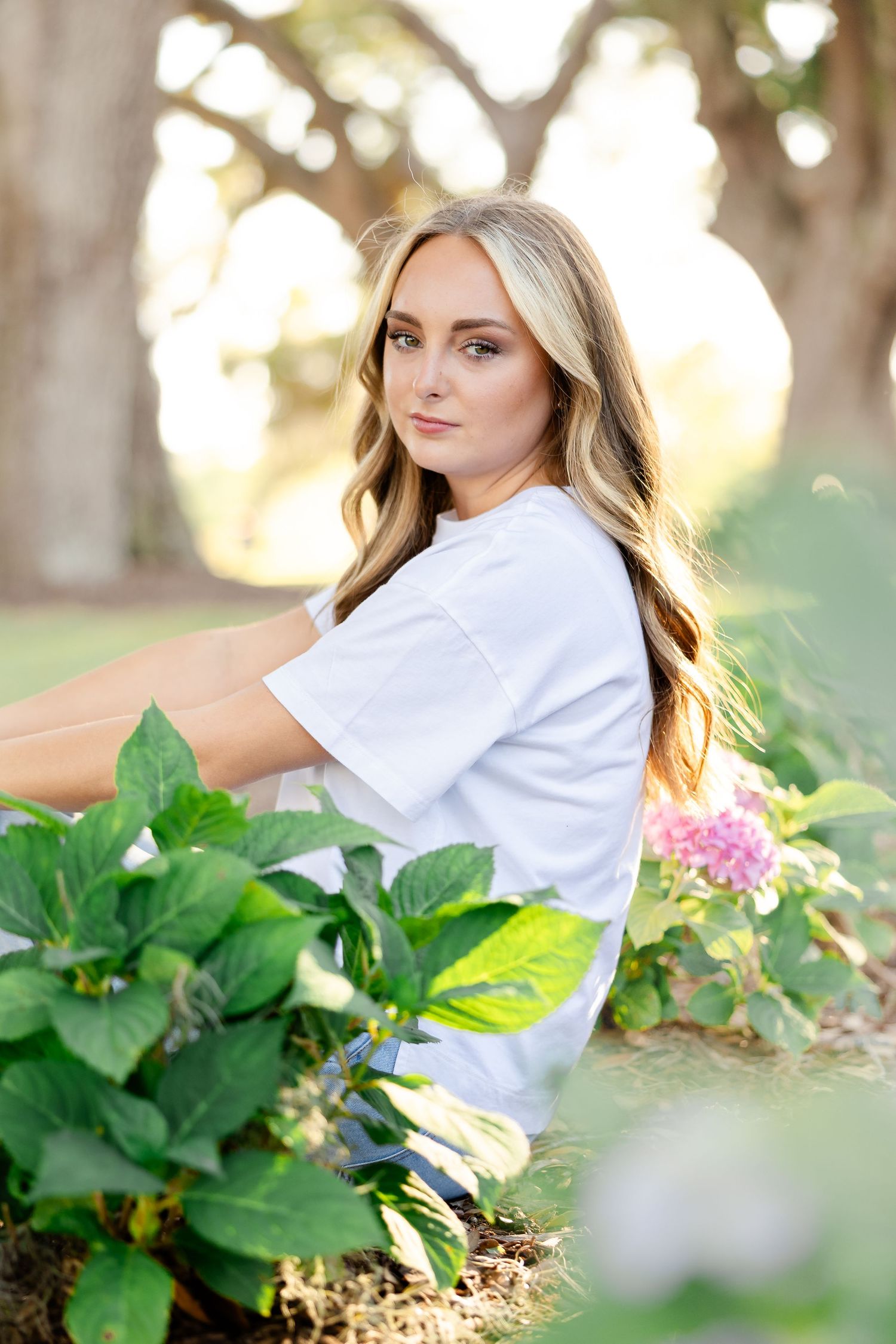 Portrait photo of someone in white t-shirt sitting by blooming hydrangeas and green plants in outdoor garden setting.