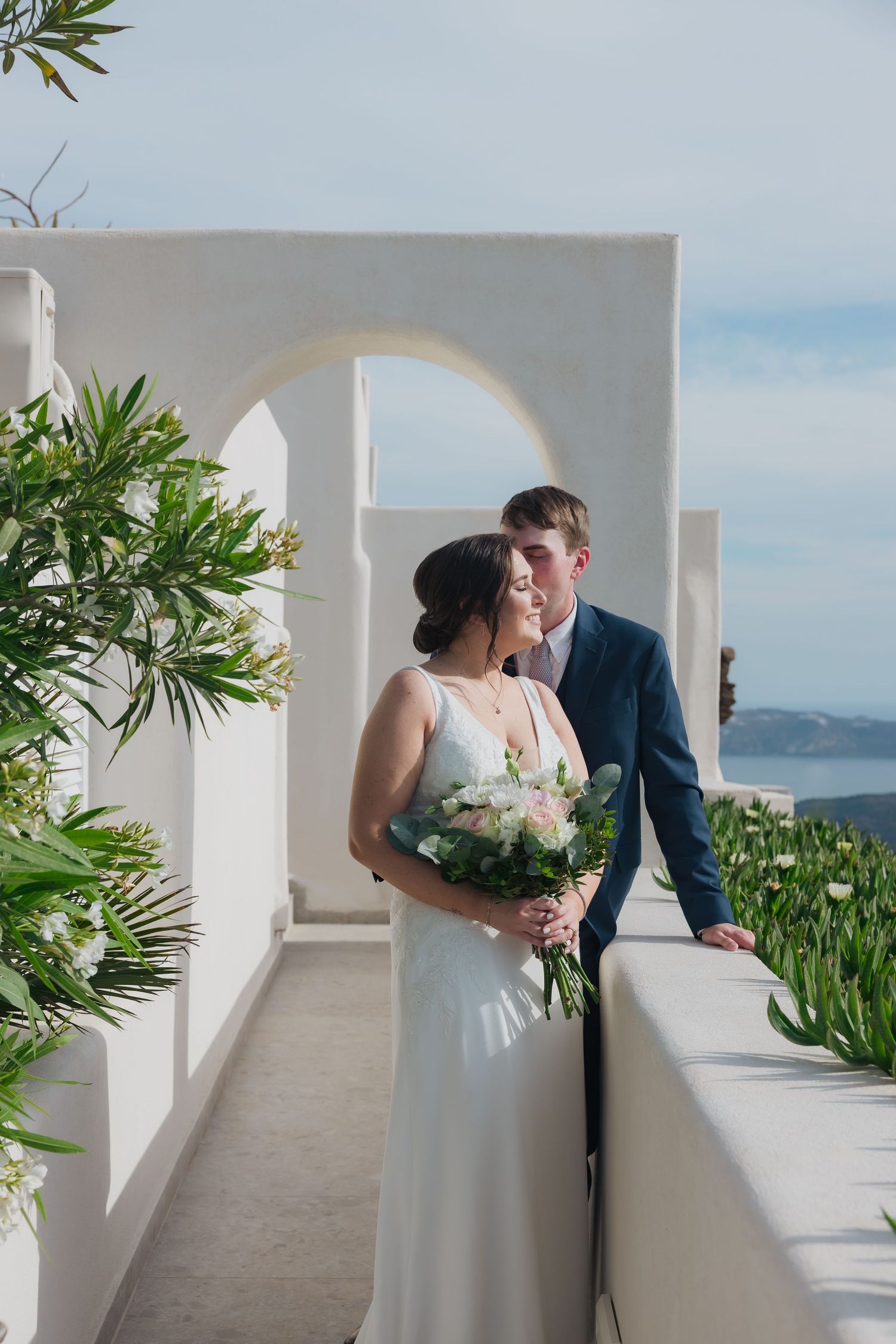 Bride and groom under white Grecian arch with lush greenery at luxury Santorini wedding venue