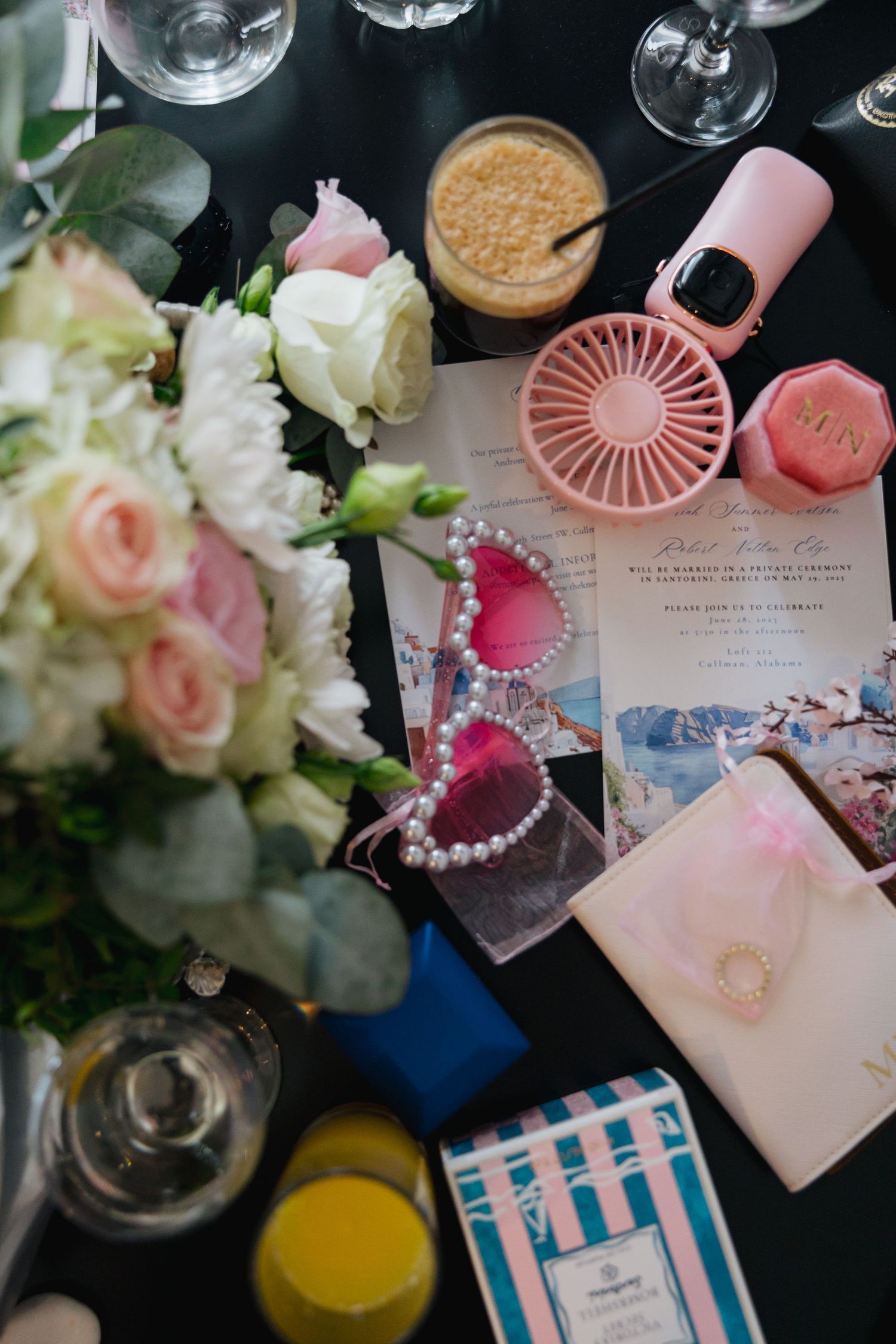 Destination wedding flat lay with pink sunglasses, bouquet, invitation, vow book, and Santorini-themed travel details