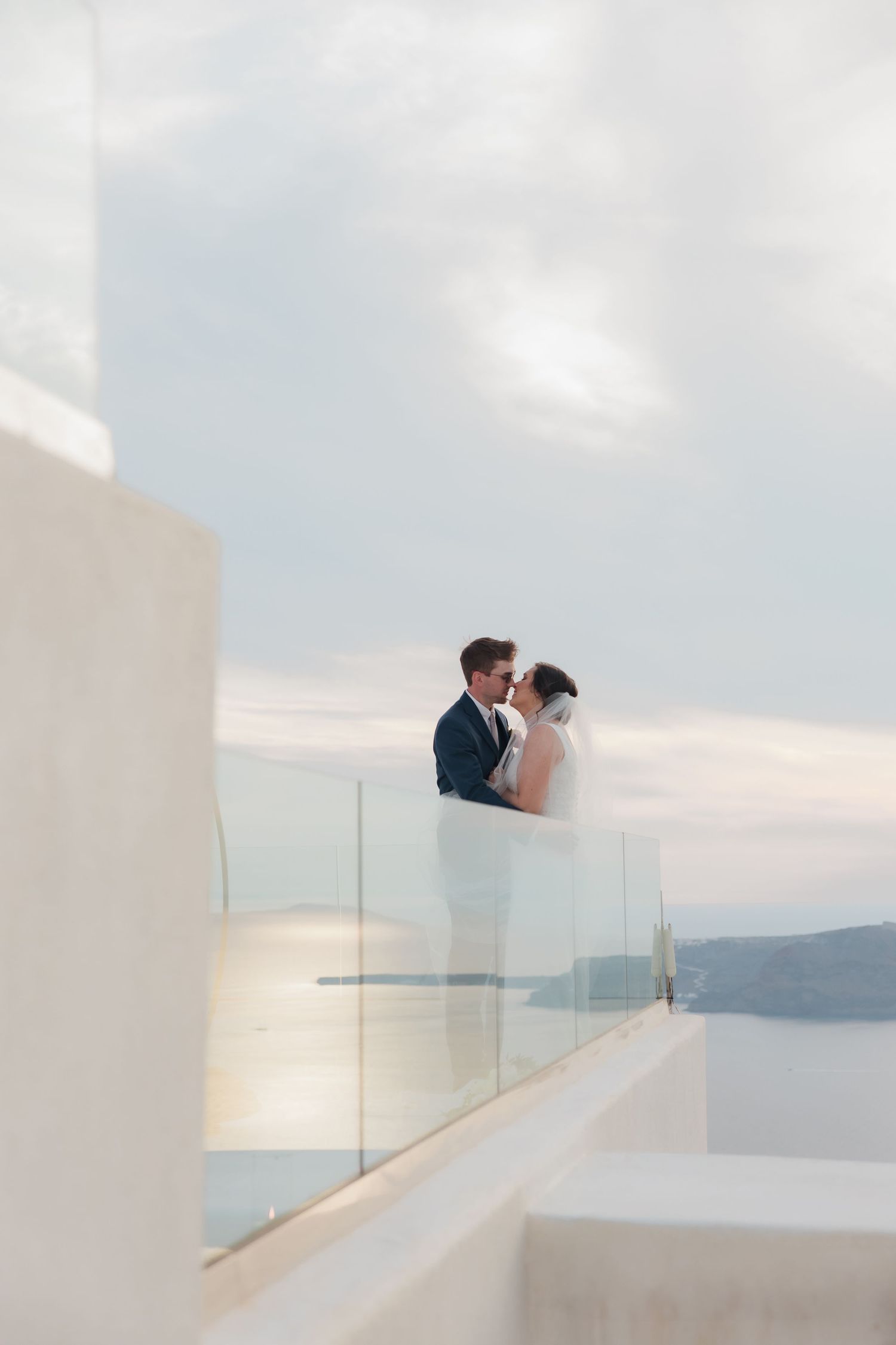 Bride and groom sharing romantic moment at sunset on glass balcony overlooking Aegean Sea