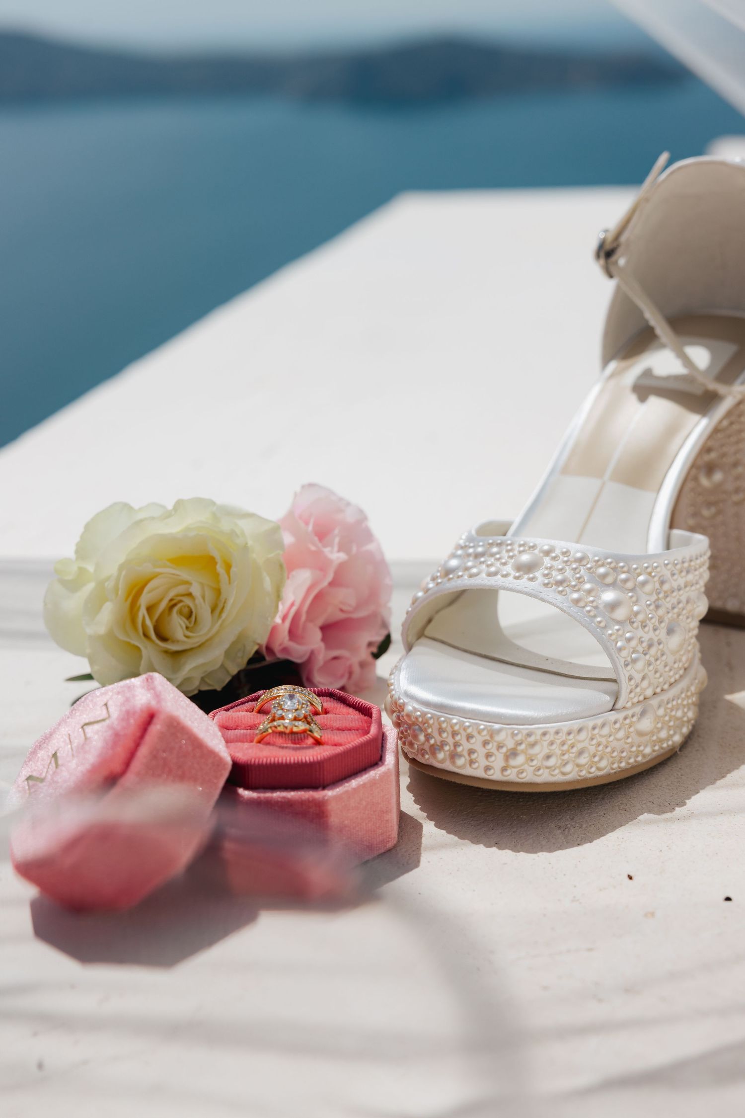 Close-up of pearl bridal heels and velvet pink ring box with yellow and blush roses at Greek island wedding