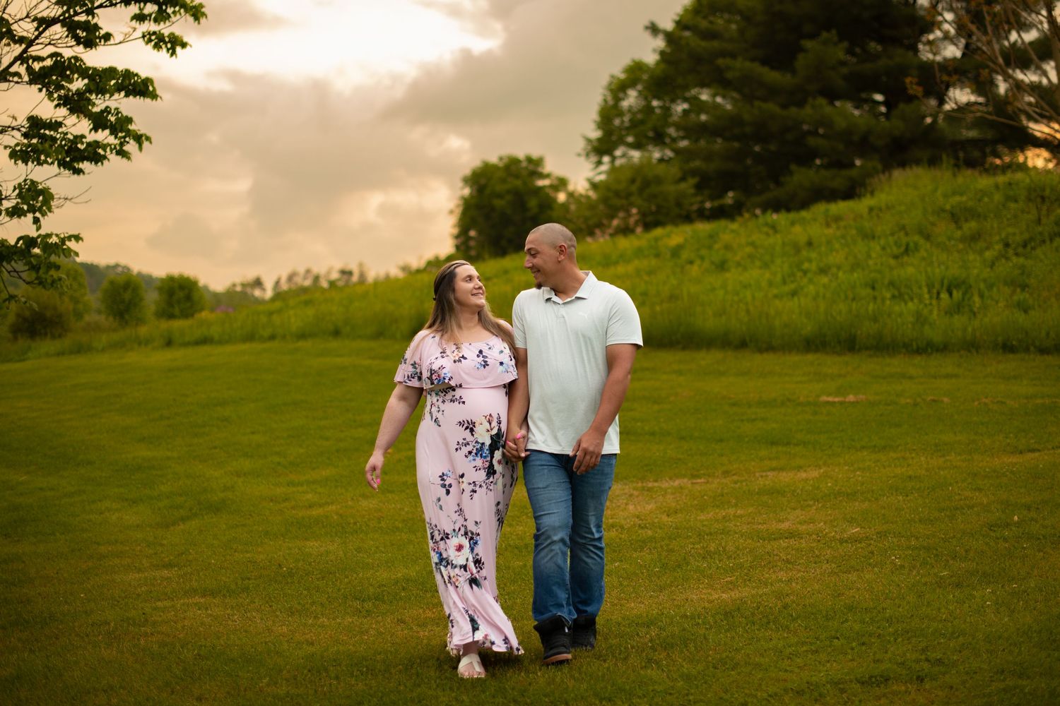 A couple walks hand in hand across a green hillside meadow during a golden sunset.
