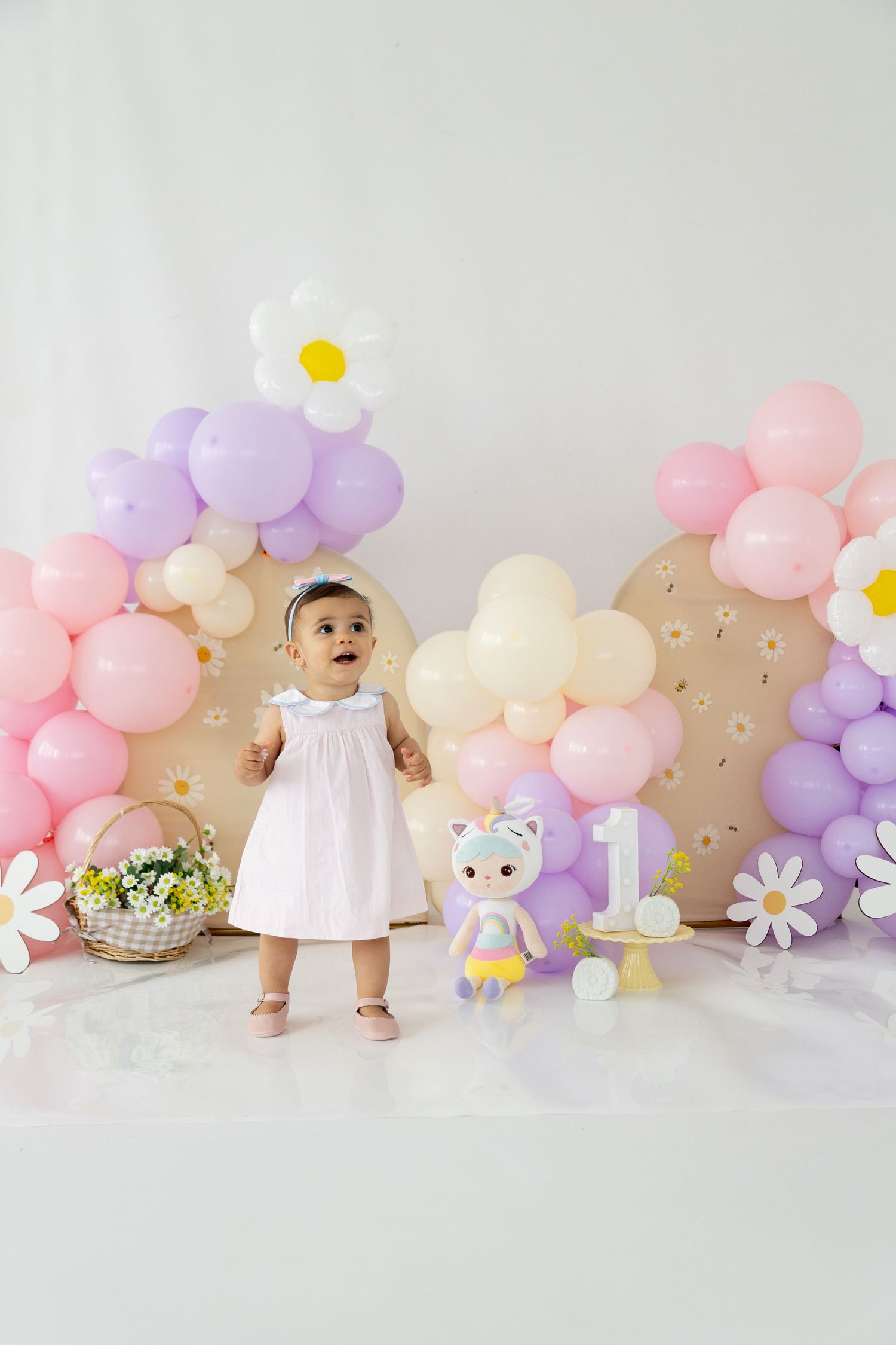 First birthday party setup with pastel balloon arch, white dress, stuffed animal and floral decor against neutral backdrop.