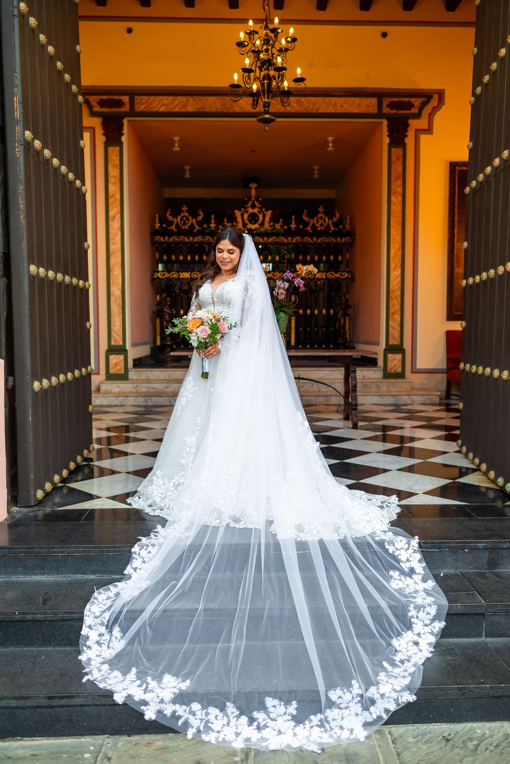 A bride in a flowing white lace dress and dramatic cathedral-length veil stands in an ornate doorway with checkered flooring.