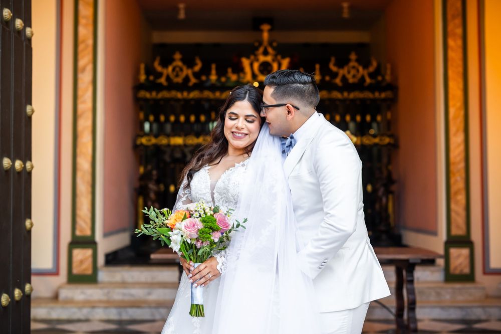 A romantic wedding couple in white attire stands in front of wine racks at a winery entrance.