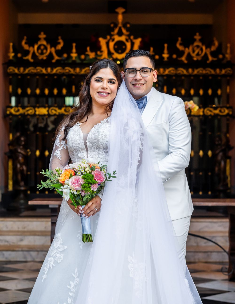 A couple in matching white wedding attire pose outside a decorative entrance with an ornate floral bouquet.