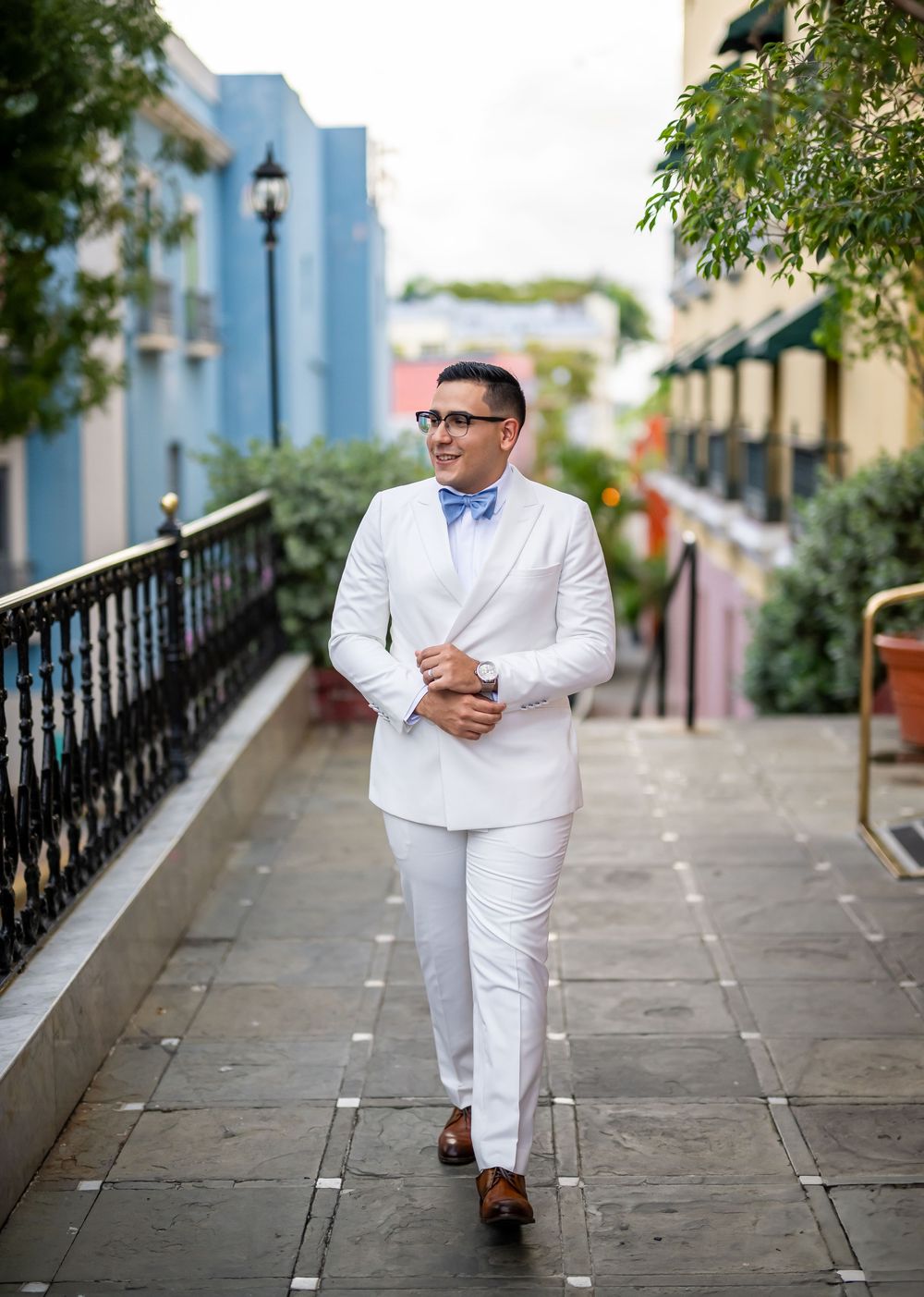 A dapper gentleman in an all-white suit and blue bow tie strolls confidently down a charming cobblestone path.
