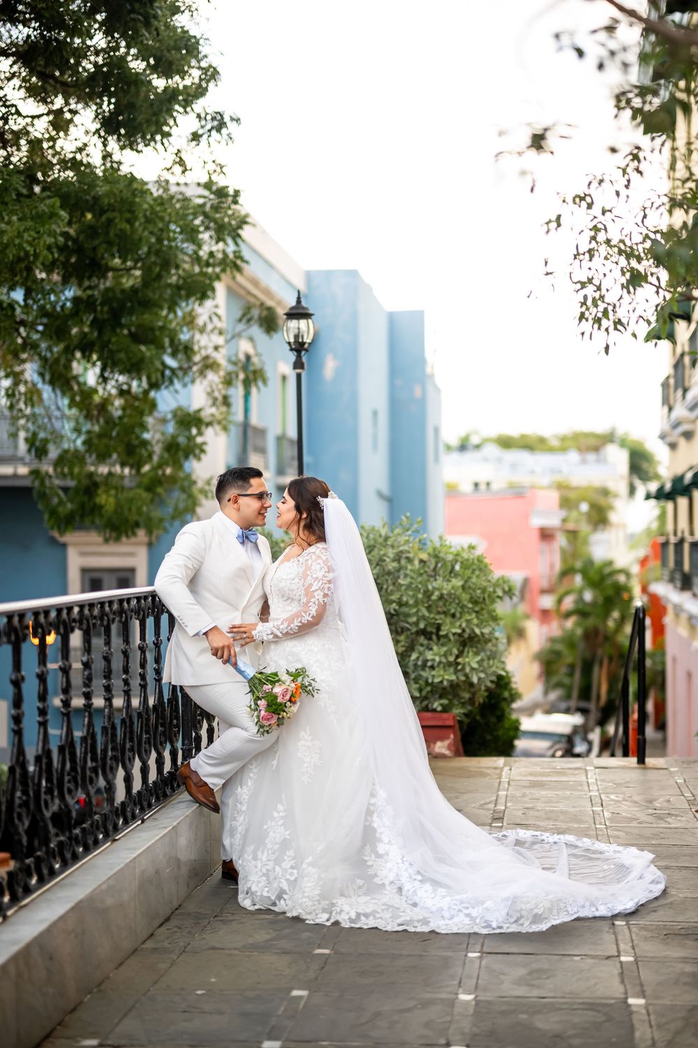 A romantic wedding couple in white poses together on stone steps with an ornate black railing and city skyline visible.