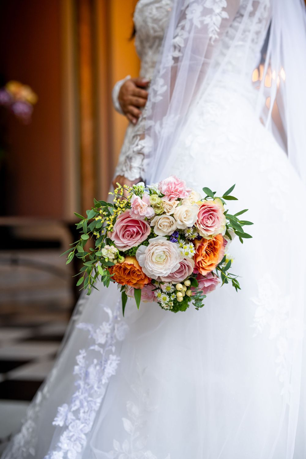 Close-up of a bridal bouquet with pink and orange roses against a white lace wedding dress.