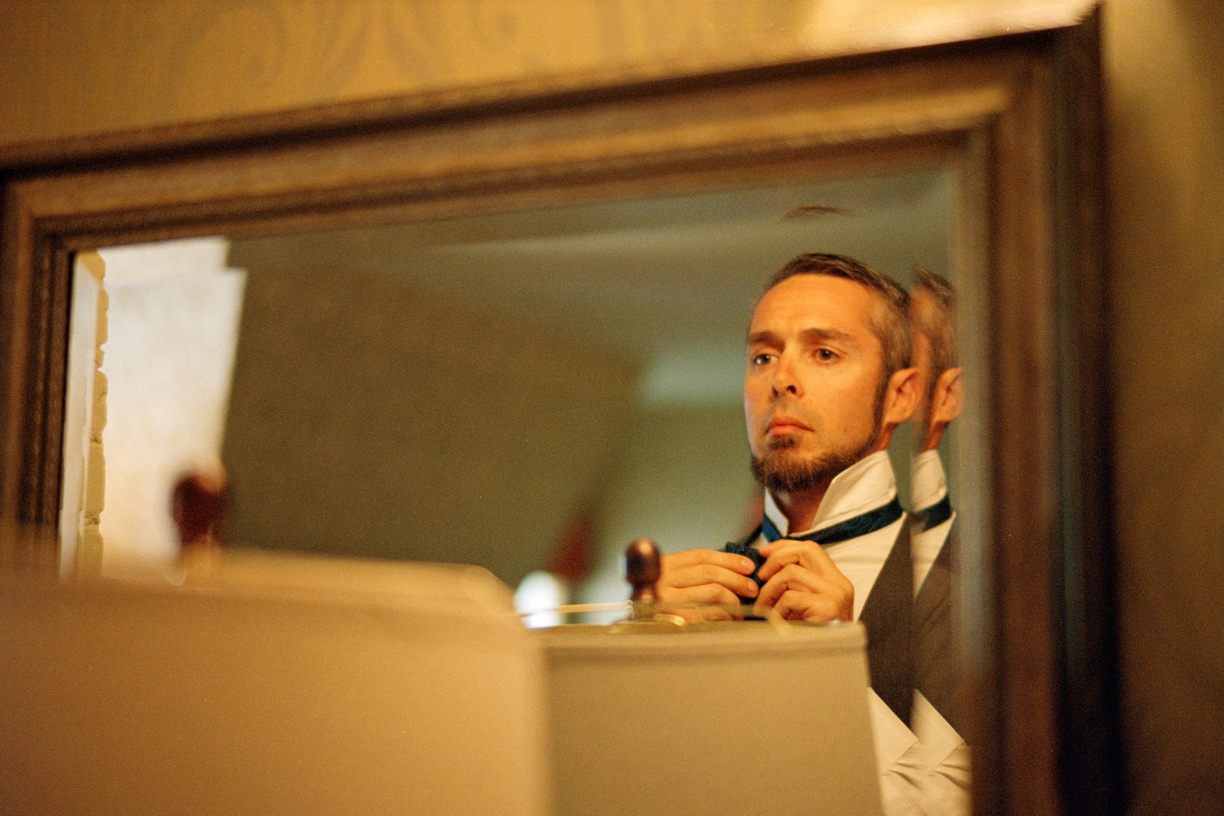 A reflection in a bathroom mirror shows someone adjusting their bowtie while getting ready.