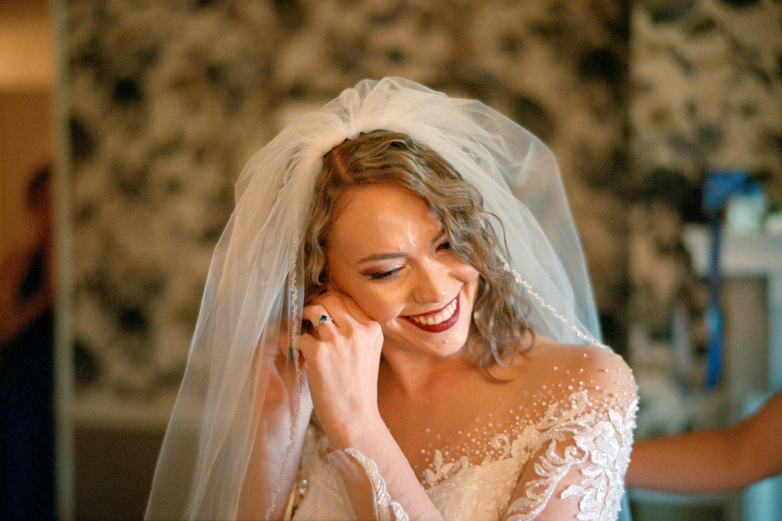 A joyful bride wearing a lace off-shoulder wedding gown and veil shares a candid moment during wedding preparations.