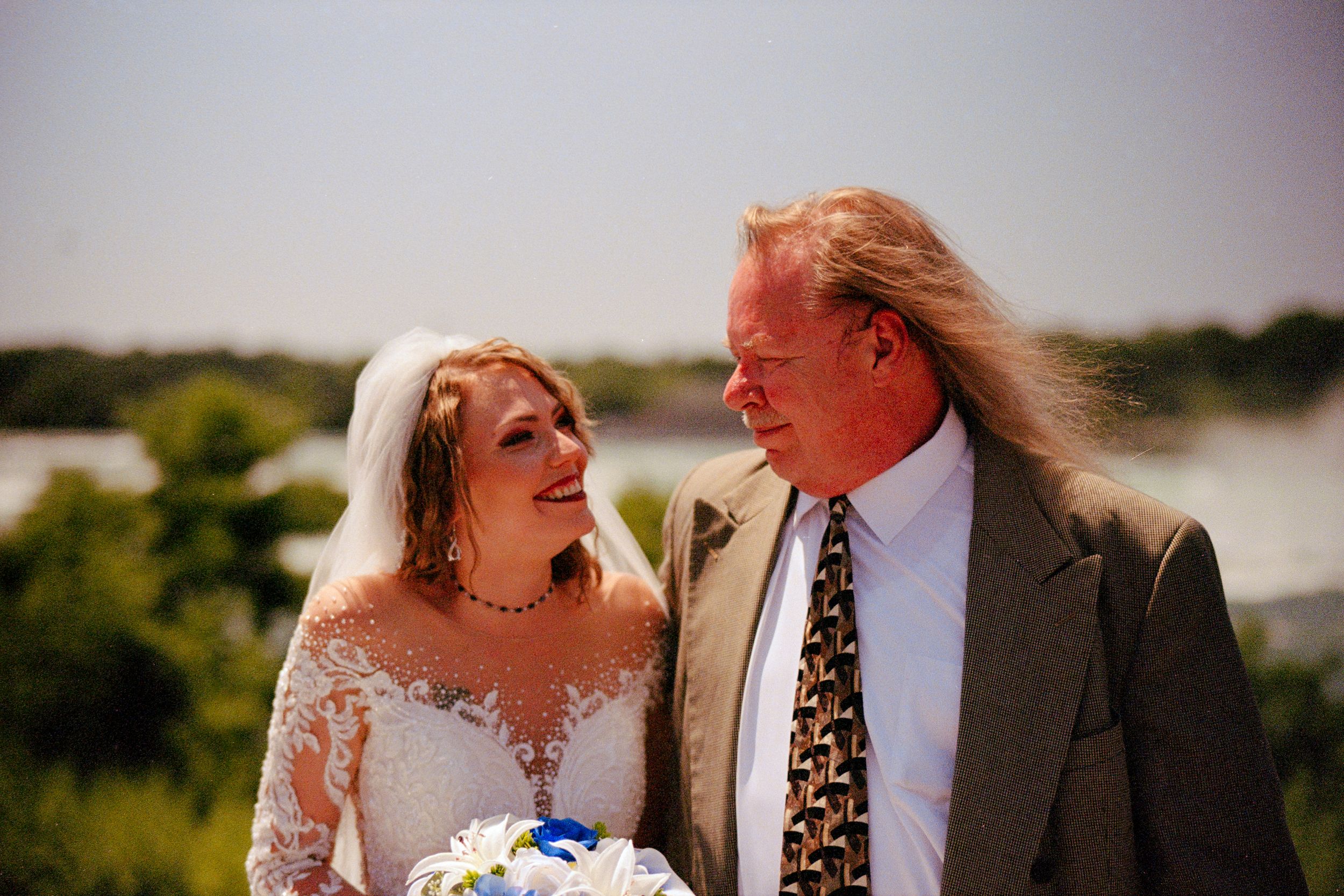 A joyful couple shares a candid moment outdoors on their wedding day with a scenic waterfront backdrop.