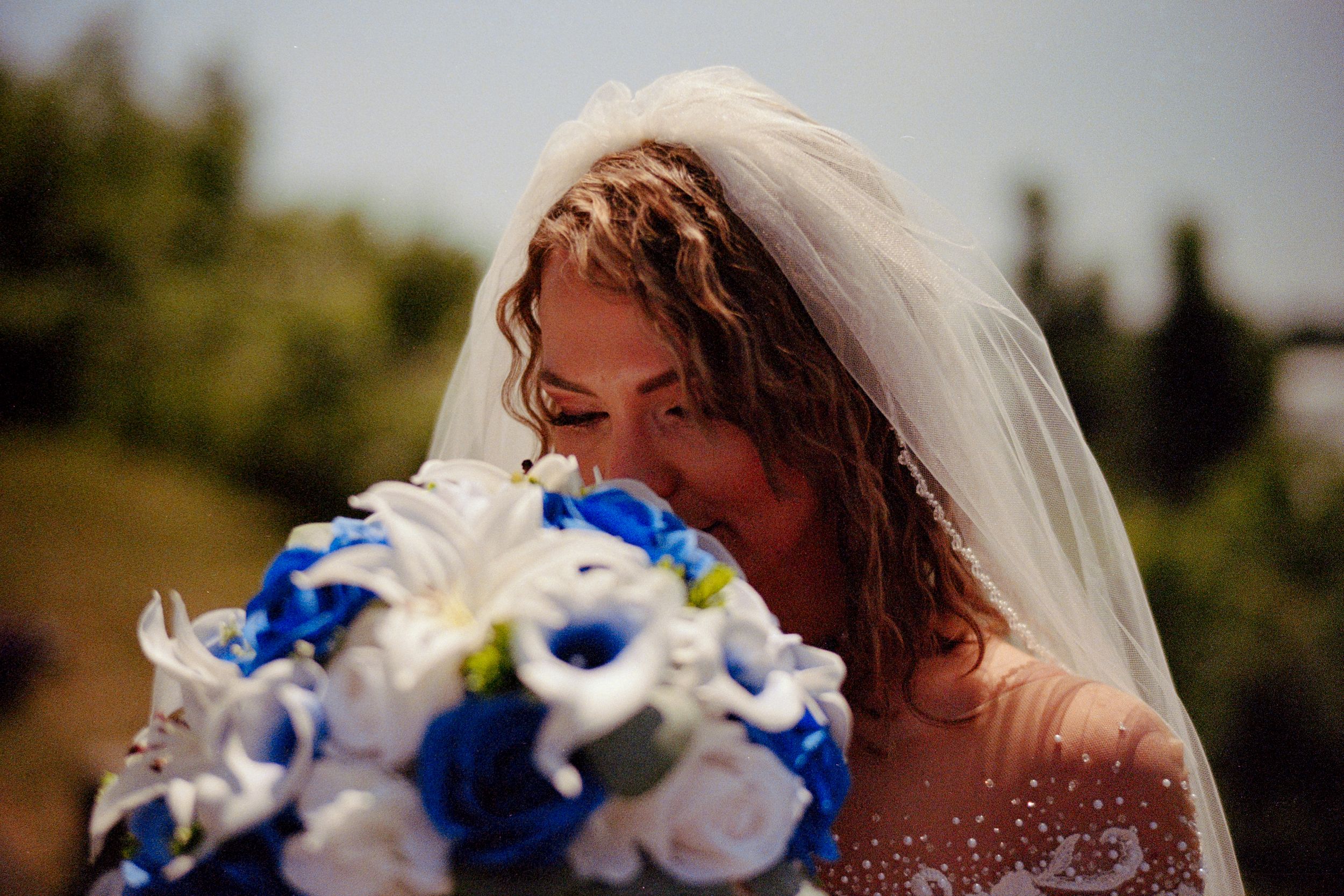 A wedding bouquet of blue and white flowers is held against a sunny outdoor background.