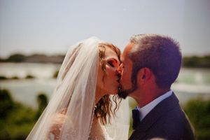 A bride and groom share a romantic kiss outdoors on their wedding day with a scenic waterfront backdrop.
