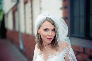 A bride in a traditional white lace wedding dress and veil poses against a brick wall background.