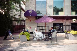 Outdoor cafe patio with purple umbrella, white chairs and yellow flowers by a modern building entrance.