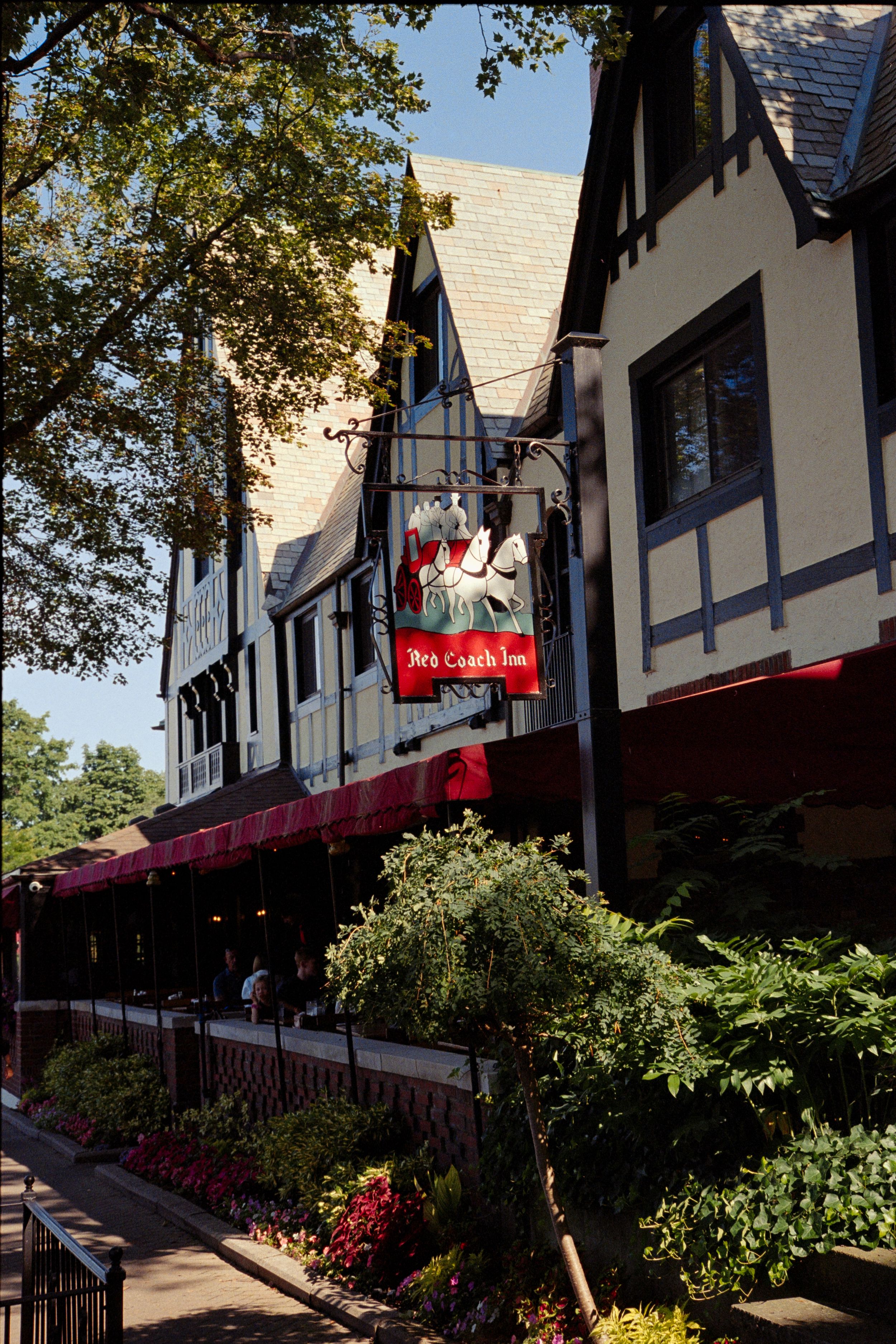 Tudor-style commercial buildings with red awnings and landscaping along a shaded sidewalk in a quaint shopping district.