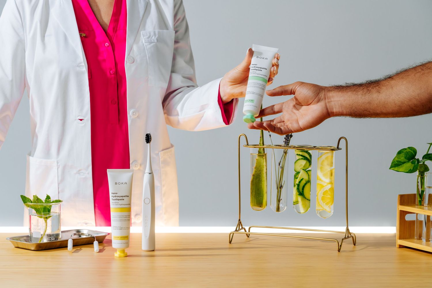 Hands exchanging skincare product with scientific equipment and plants on wooden table.