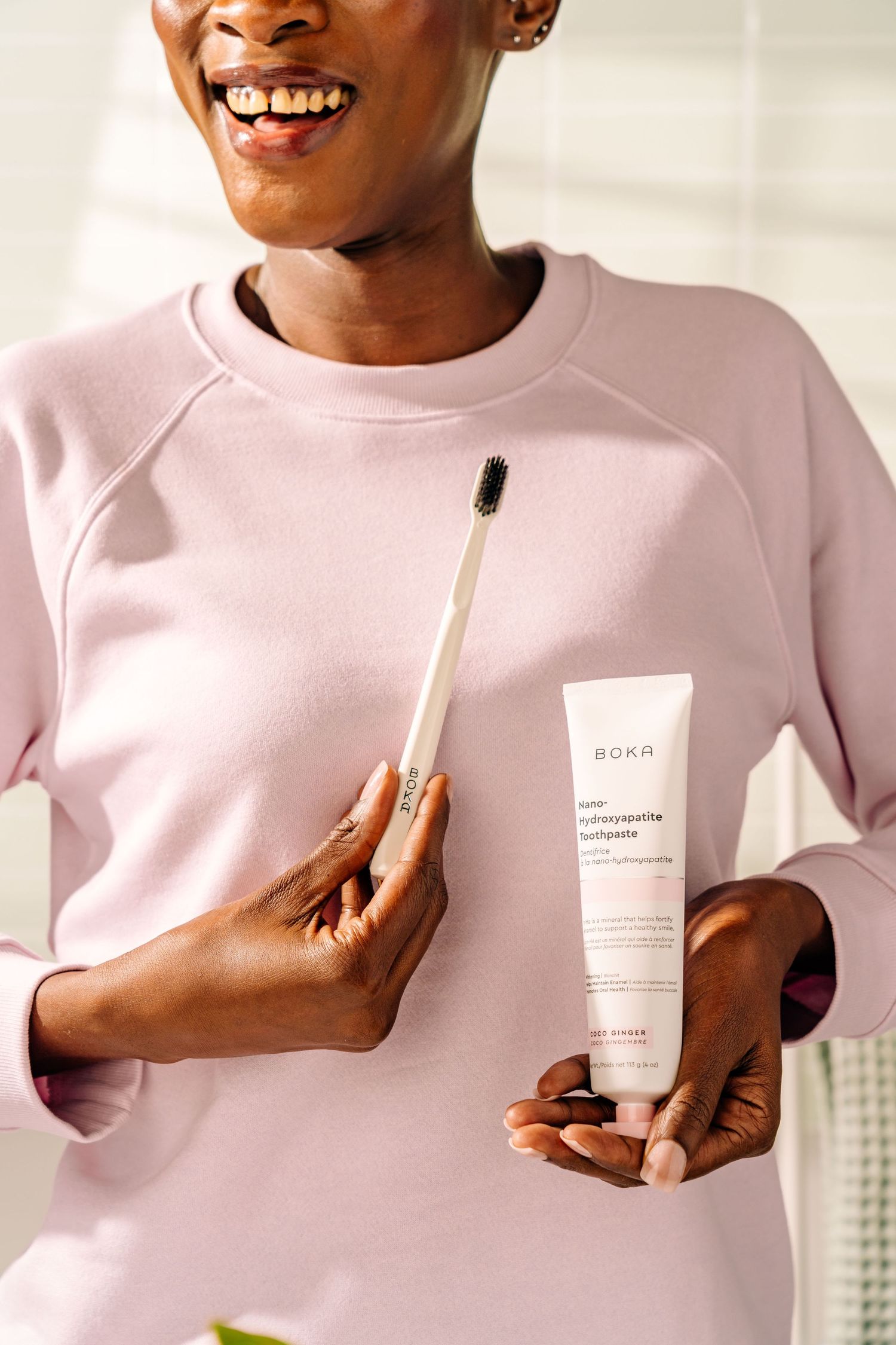 Person wearing pink sweatshirt holding bamboo toothbrush and natural beauty product tube against white background.