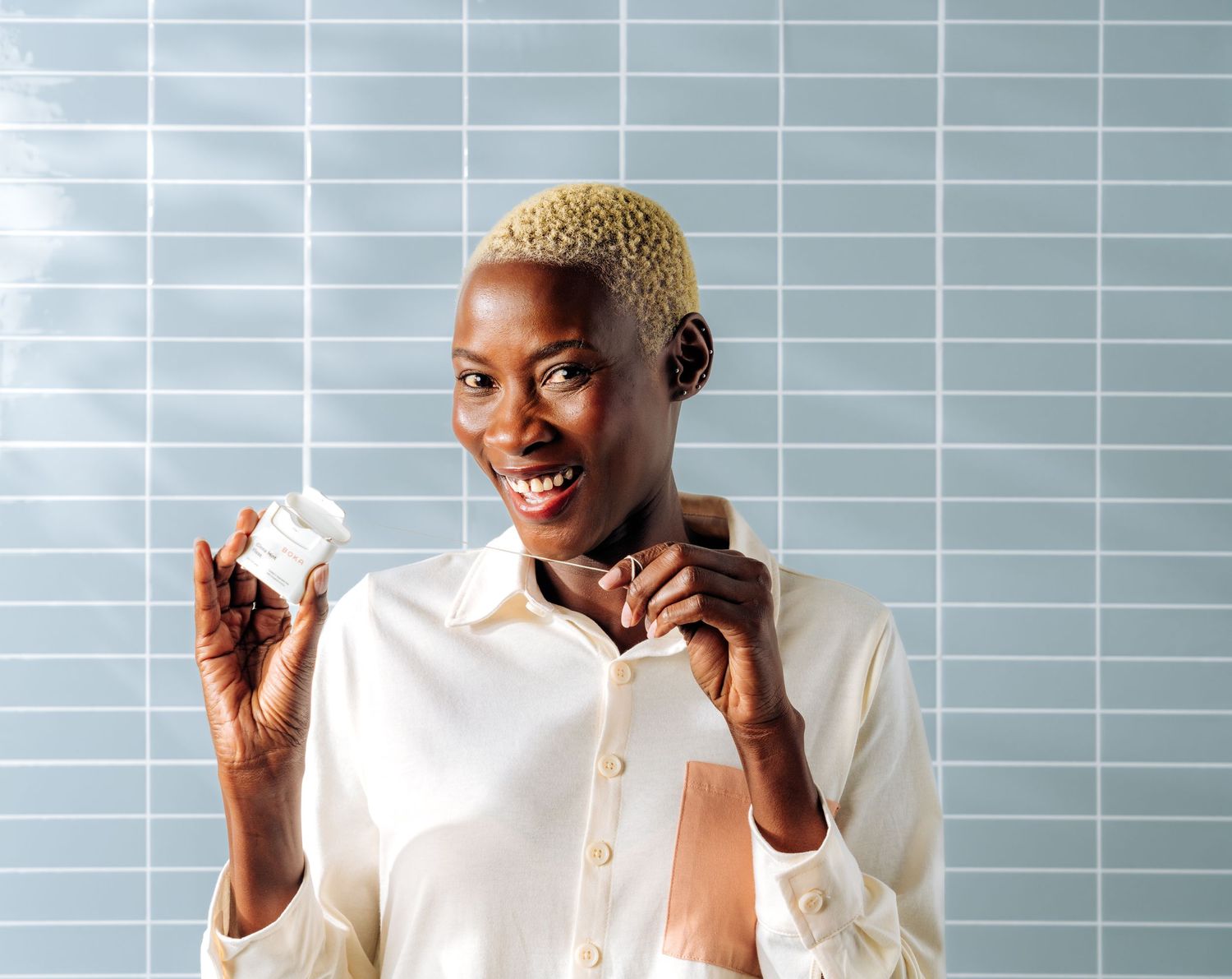 Person wearing white shirt holding a small skincare product against a blue tiled wall background.