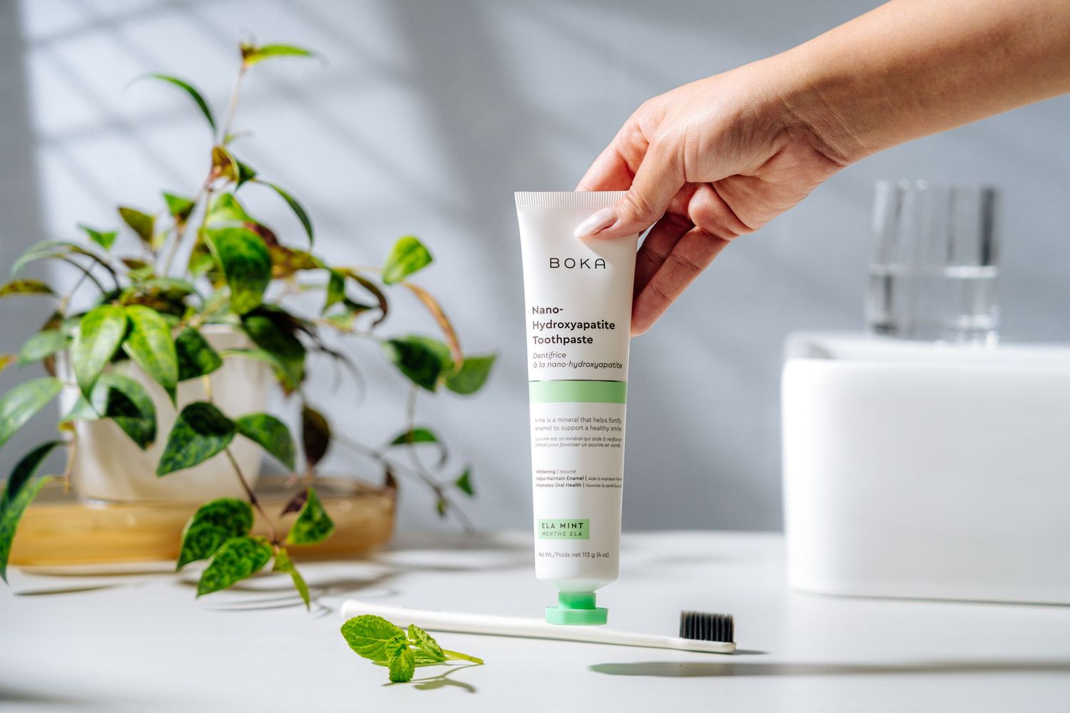 Hand holding a white skincare product tube against a bathroom counter with green plants.