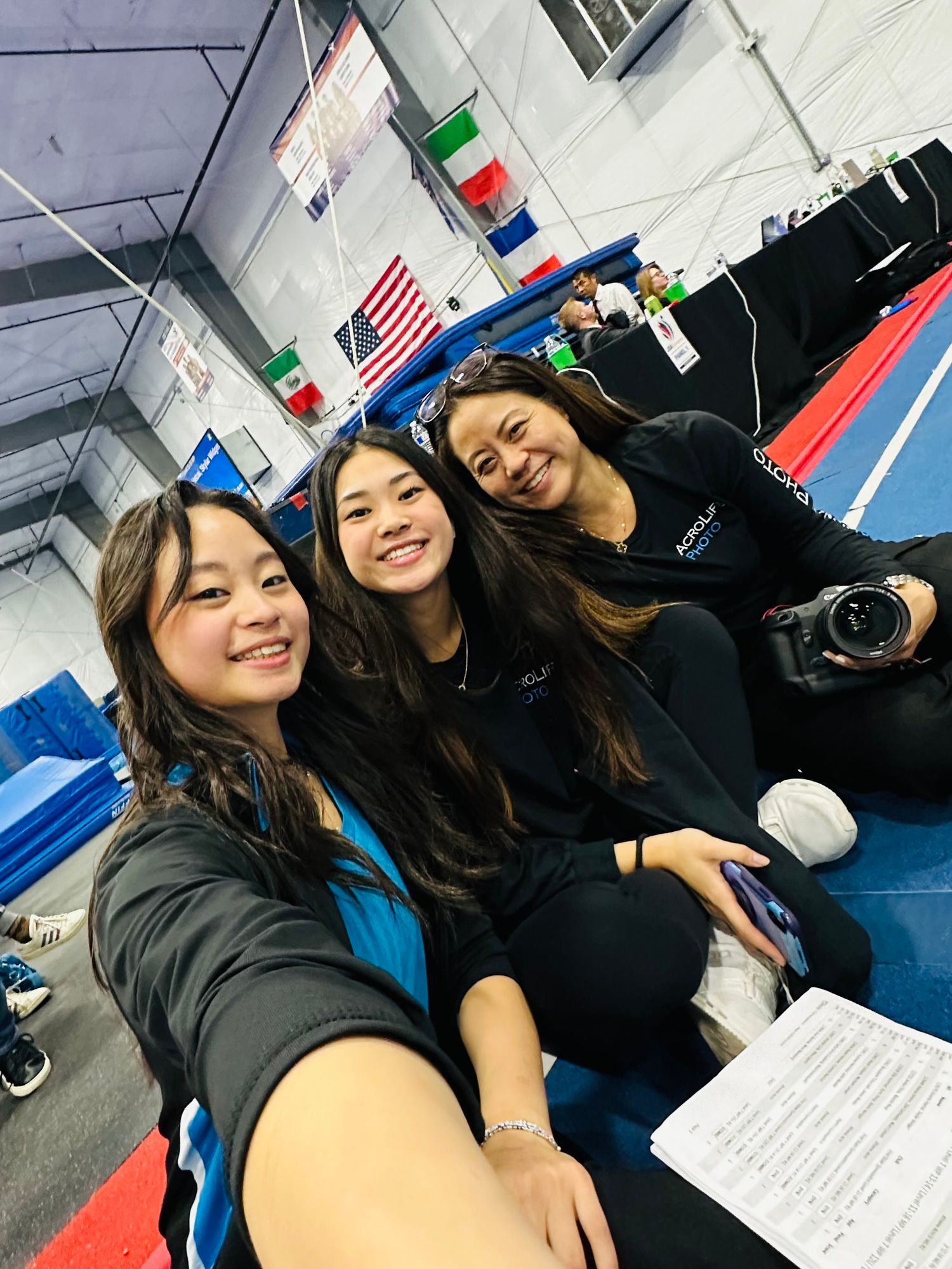 Three teammates take selfie in gymnastics facility with flags hanging in background.