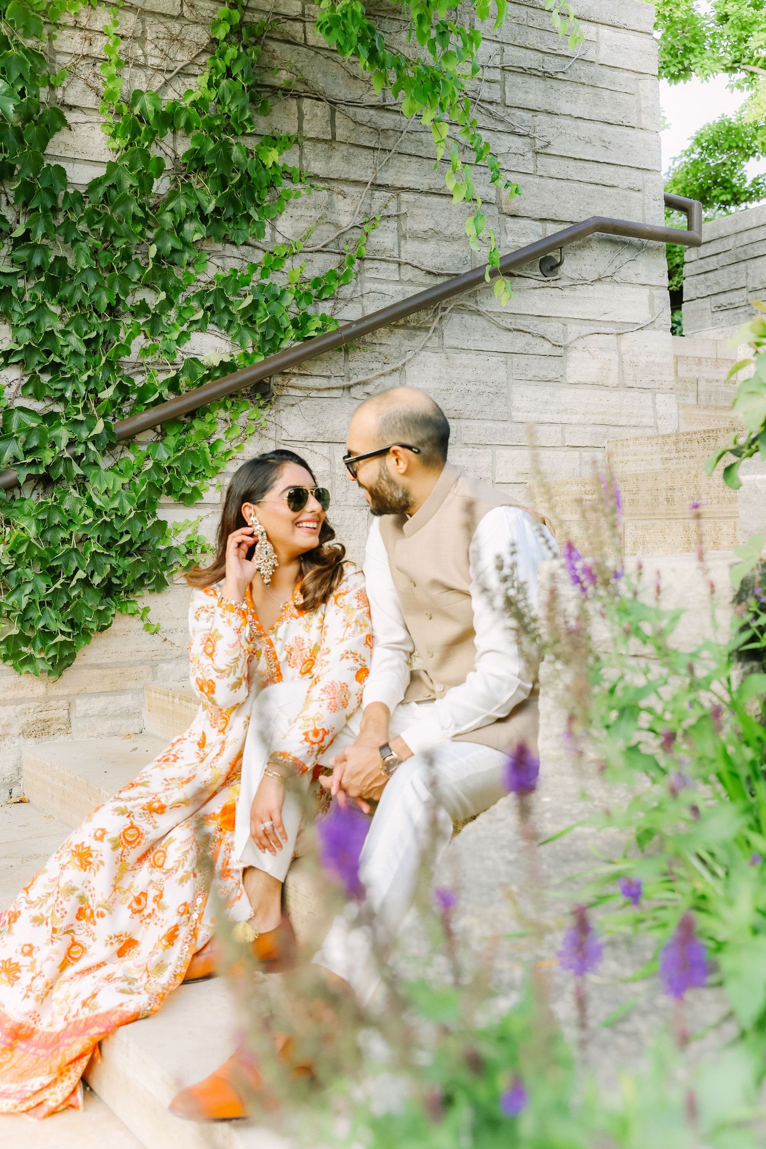 South Asian couple in traditional attire sitting on stone steps surrounded by lush greenery and flowers.