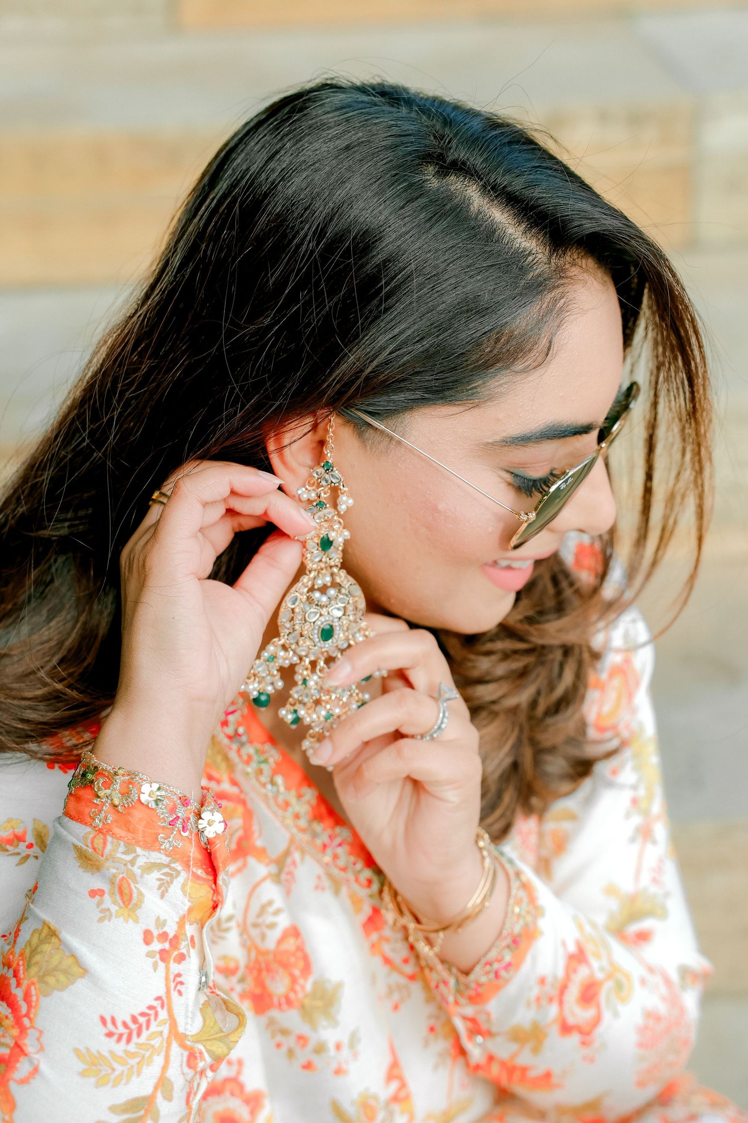 South Asian bride in orange floral outfit adjusting ornate emerald and diamond earring with sunglasses.