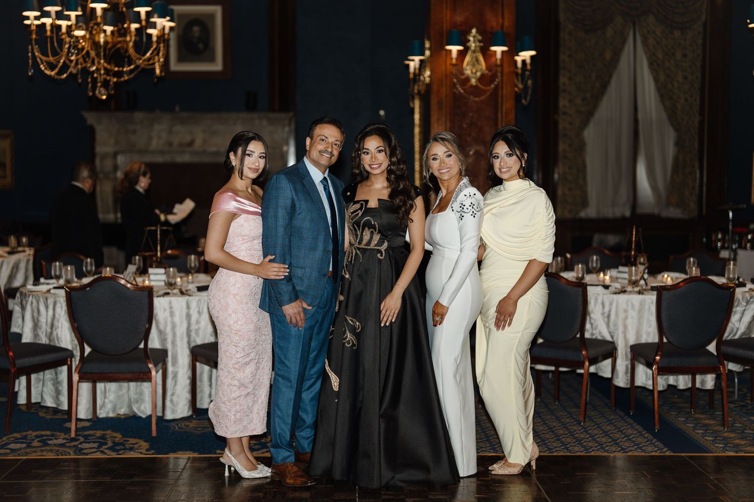 Group of five elegantly dressed people posing together in an ornate blue-walled event venue.
